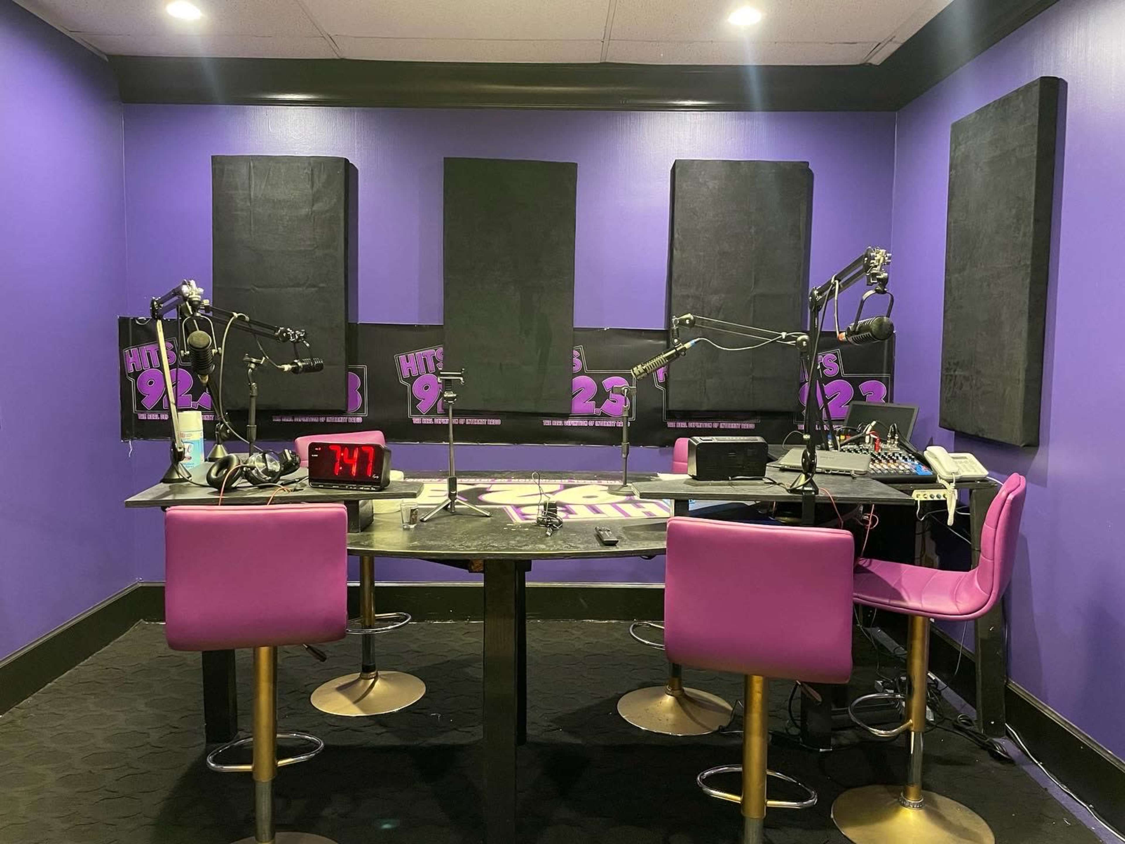 A radio studio with purple walls, two pink chairs, and equipment including microphones and a mixer on a central table.