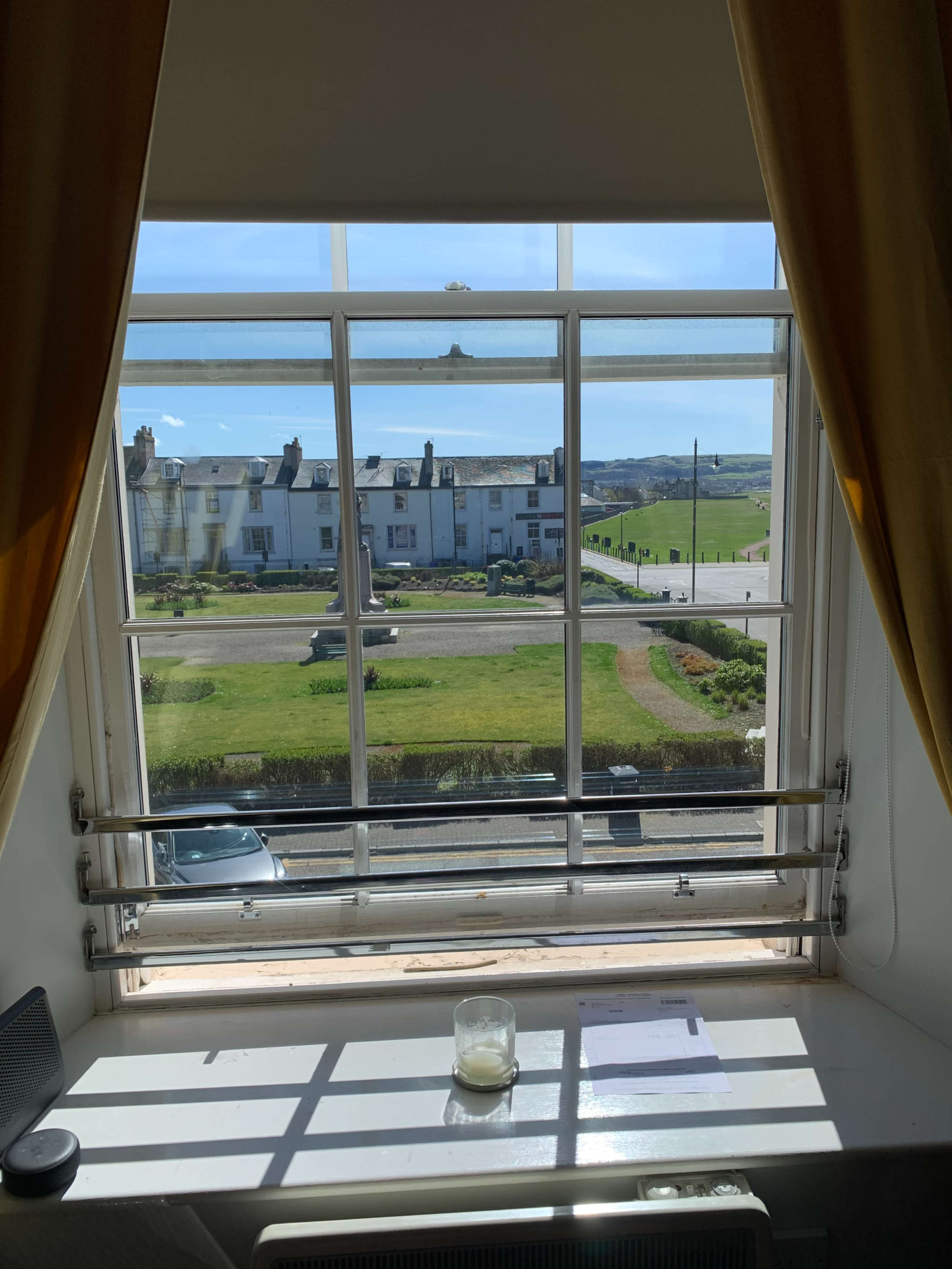 A window frame provides a view of a street lined with houses and green fields under a clear sky.