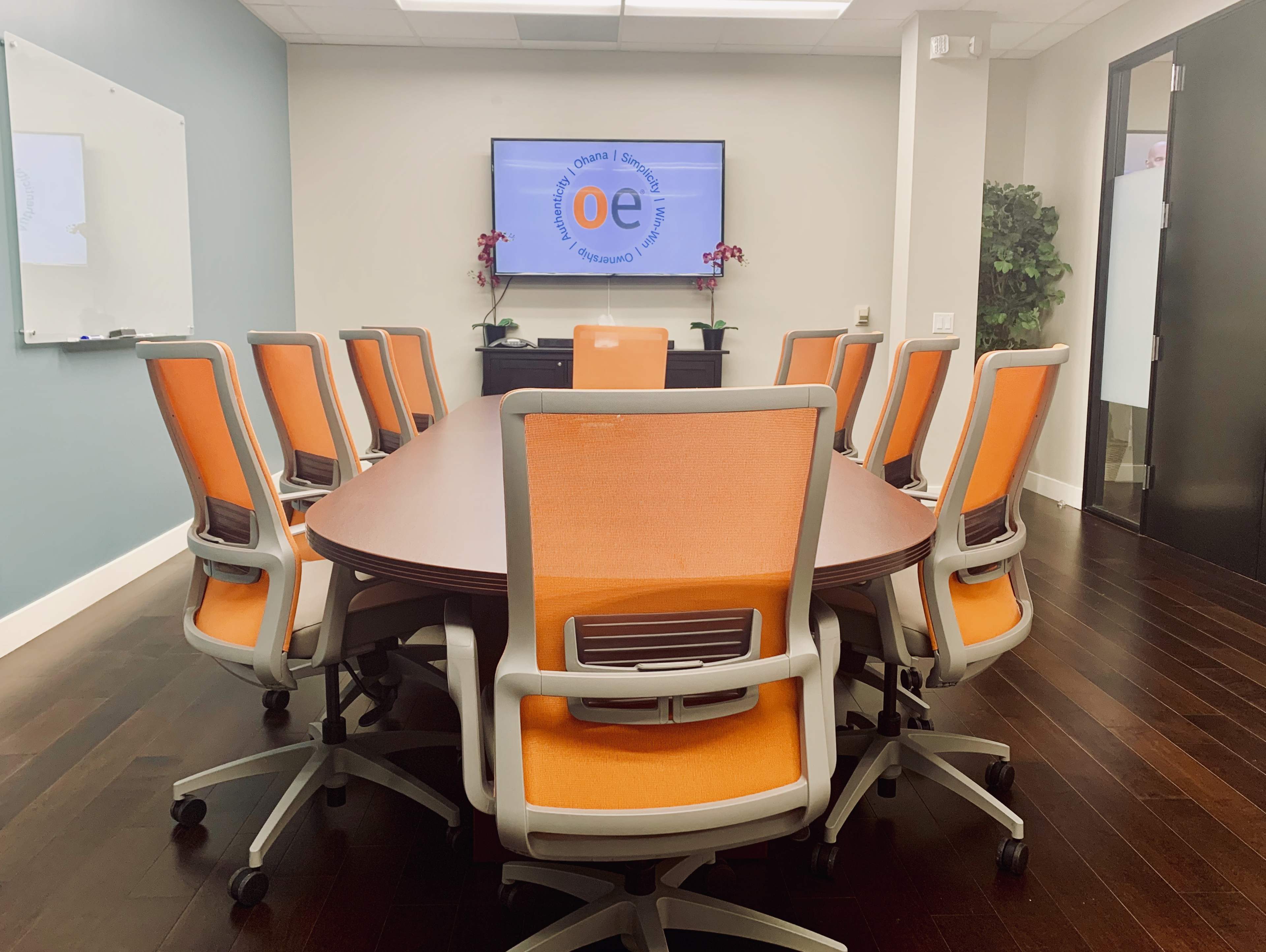 A conference room features a large oval table surrounded by ten orange and gray chairs, with a television displaying a logo on the wall.
