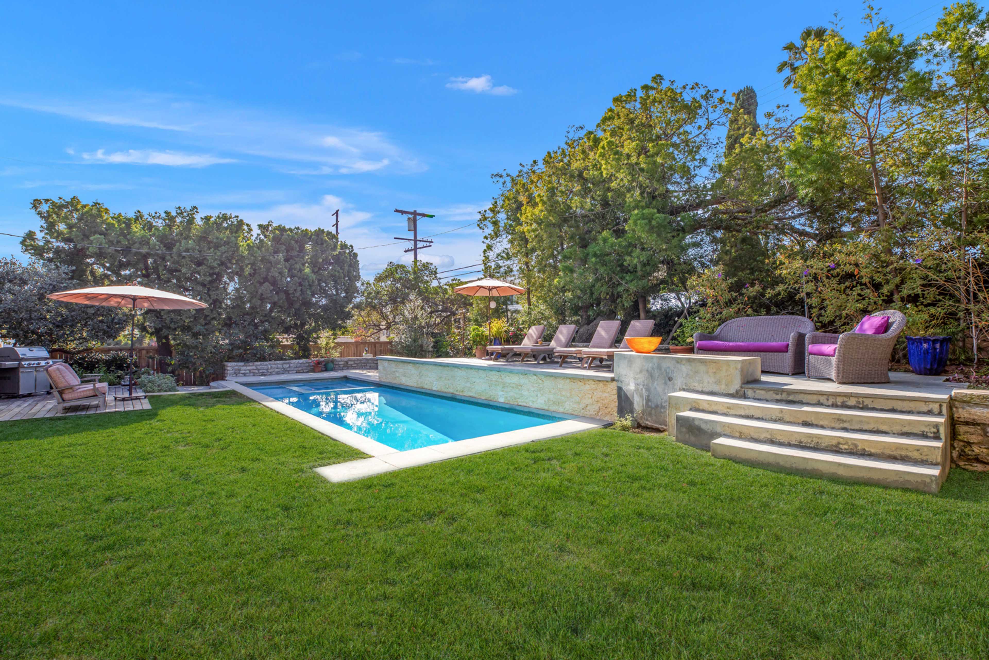 The image shows a modern backyard featuring a blue swimming pool surrounded by green grass, several lounge chairs, and two large umbrellas.