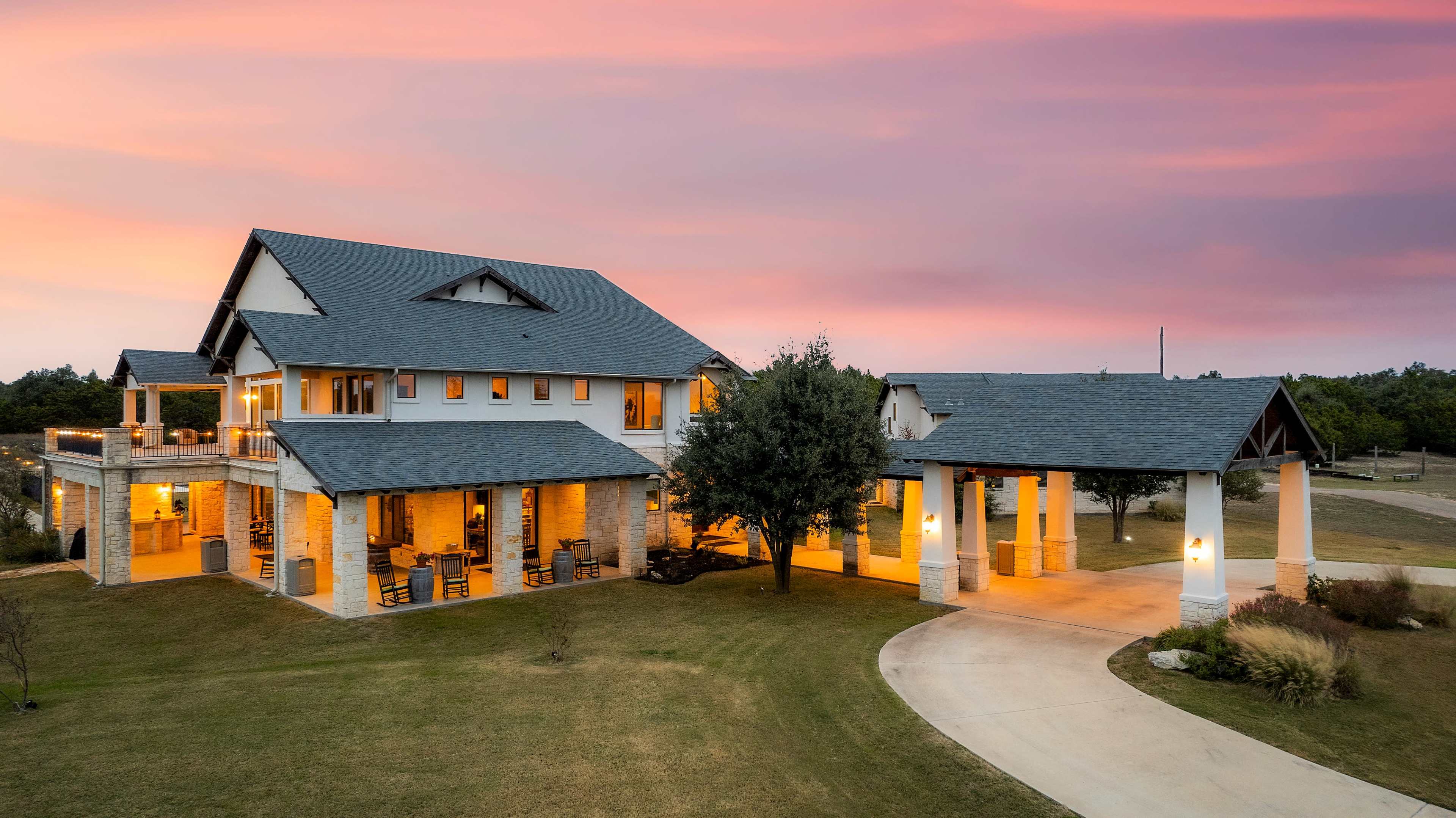 A modern two-story house with a stone exterior and large windows sits at dusk, illuminated by warm lights under a colorful sky.