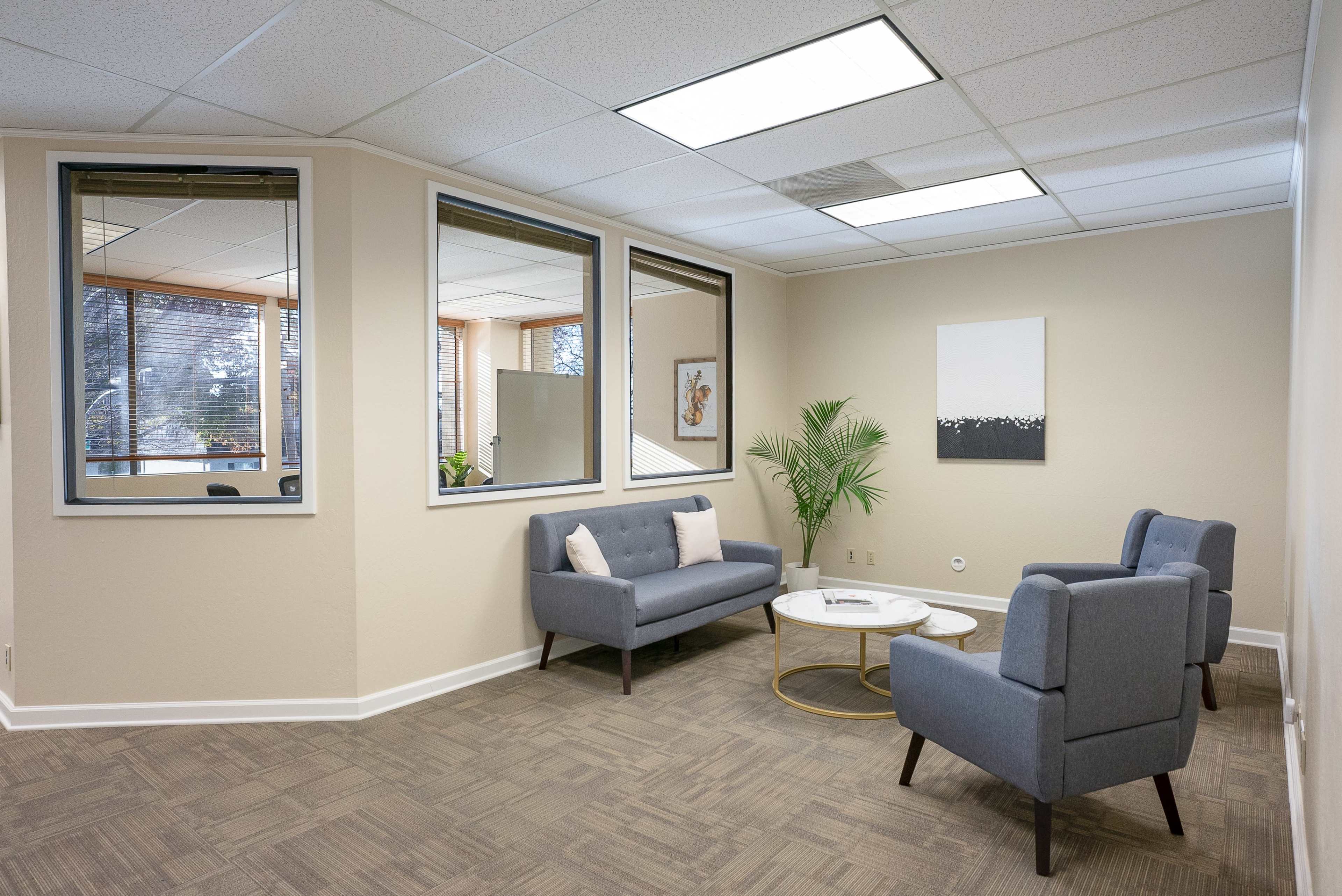 The image displays a modern office waiting area featuring two gray sofas, a round coffee table, and large windows allowing natural light to illuminate the space.