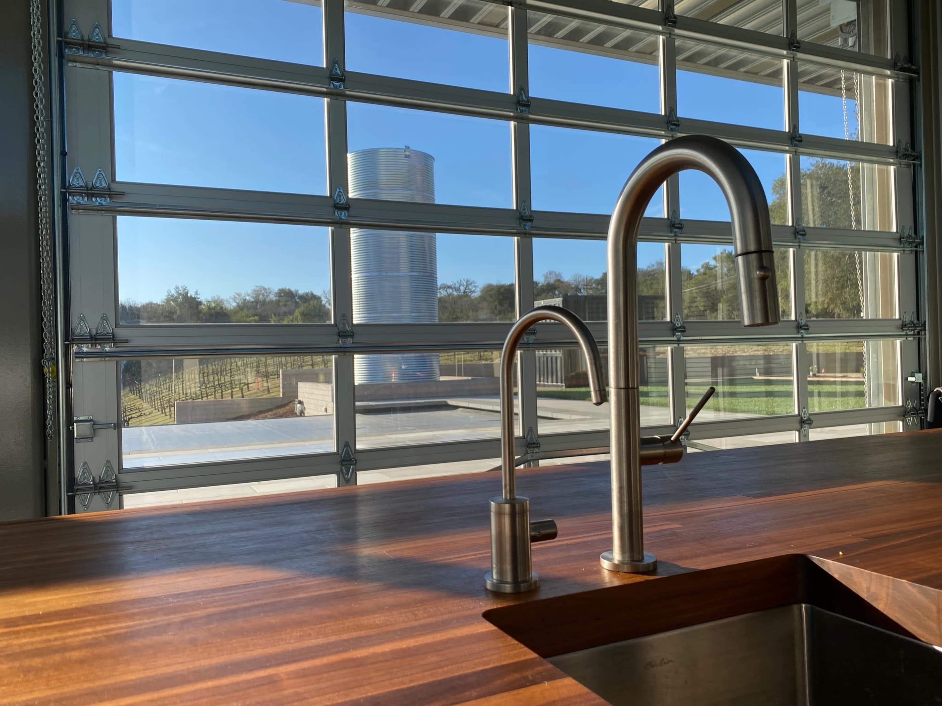 A modern kitchen features a stainless steel faucet in the foreground, with large glass garage doors showing an outdoor view of a metal silo and green landscape beyond.