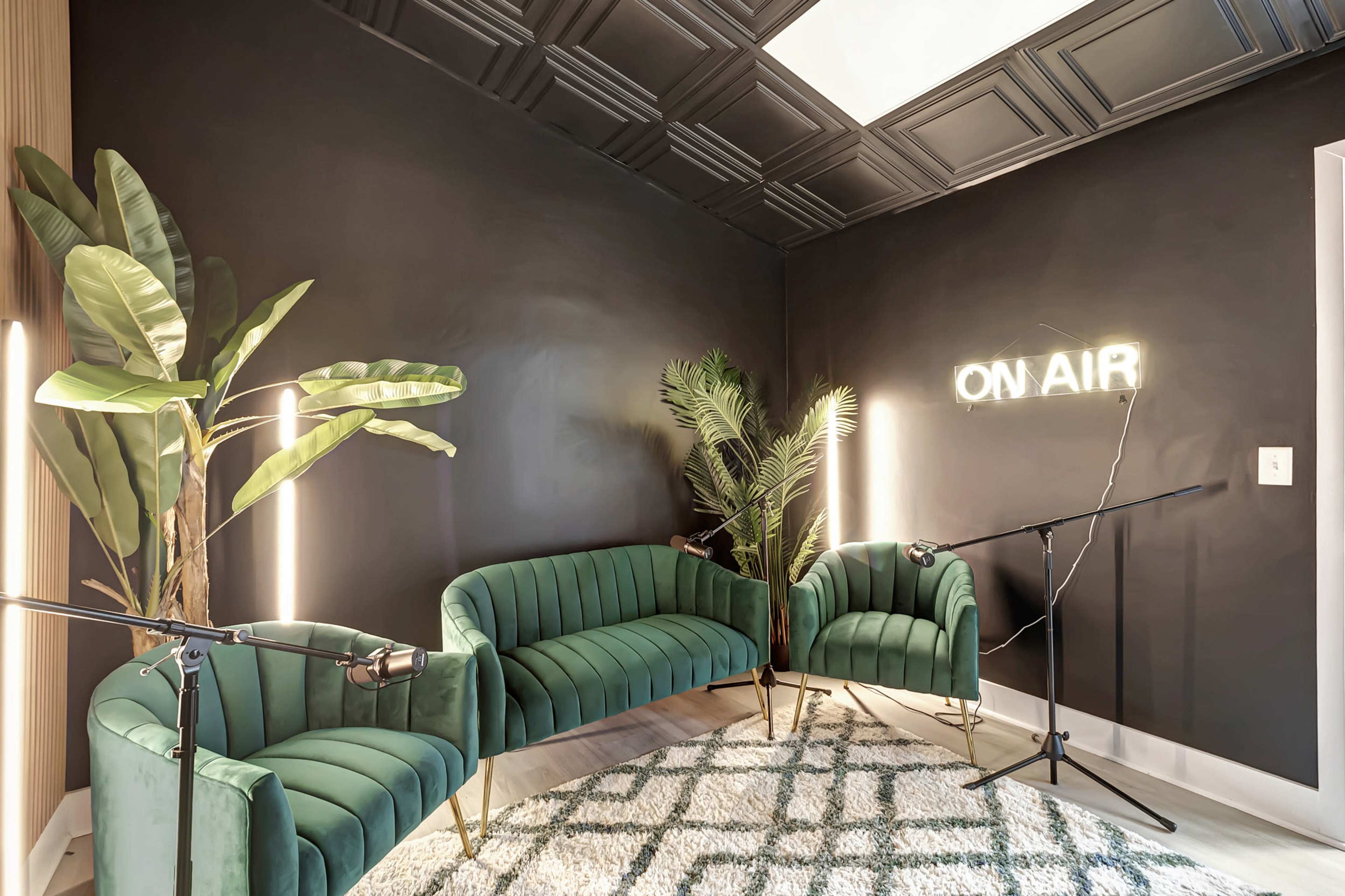 A small recording studio features green velvet chairs, a patterned rug, large plants, and an illuminated "ON AIR" sign against a black wall.