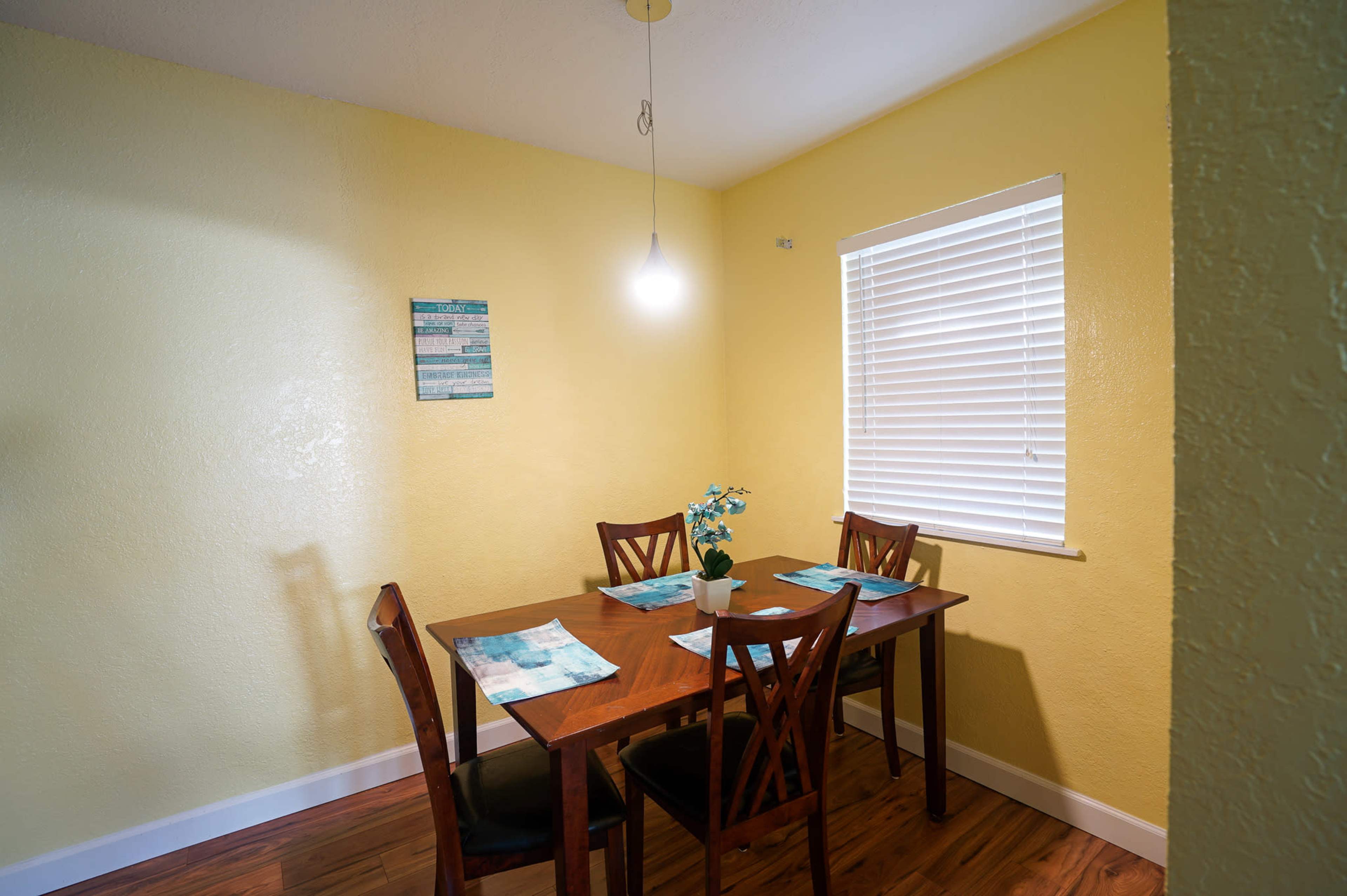 A small dining area features a wooden table with four chairs, a potted plant, and a window with blinds.