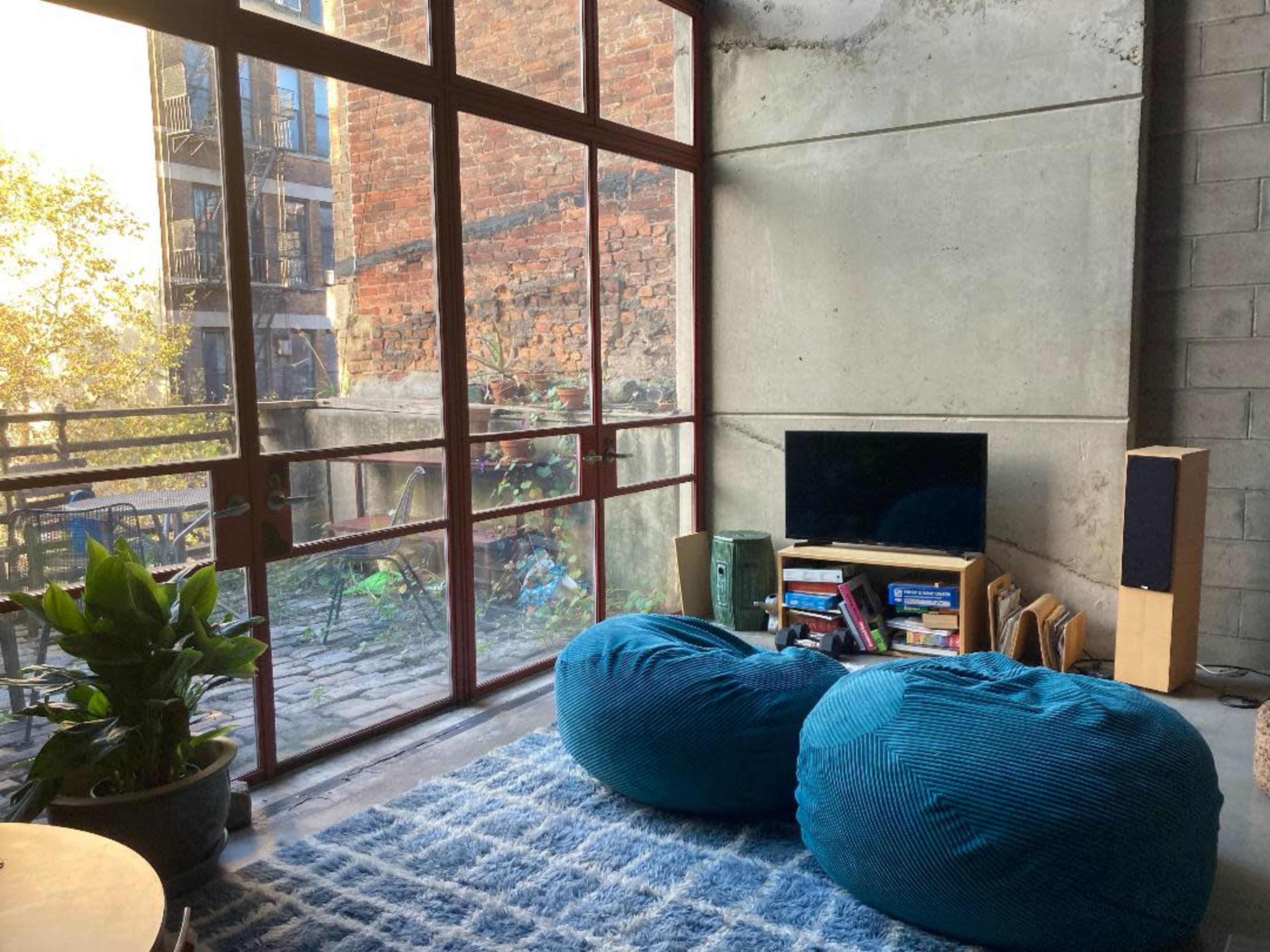 The scene features a modern living space with two blue bean bag chairs, a television on a wooden stand, a plant, and large glass windows revealing a brick exterior outside.