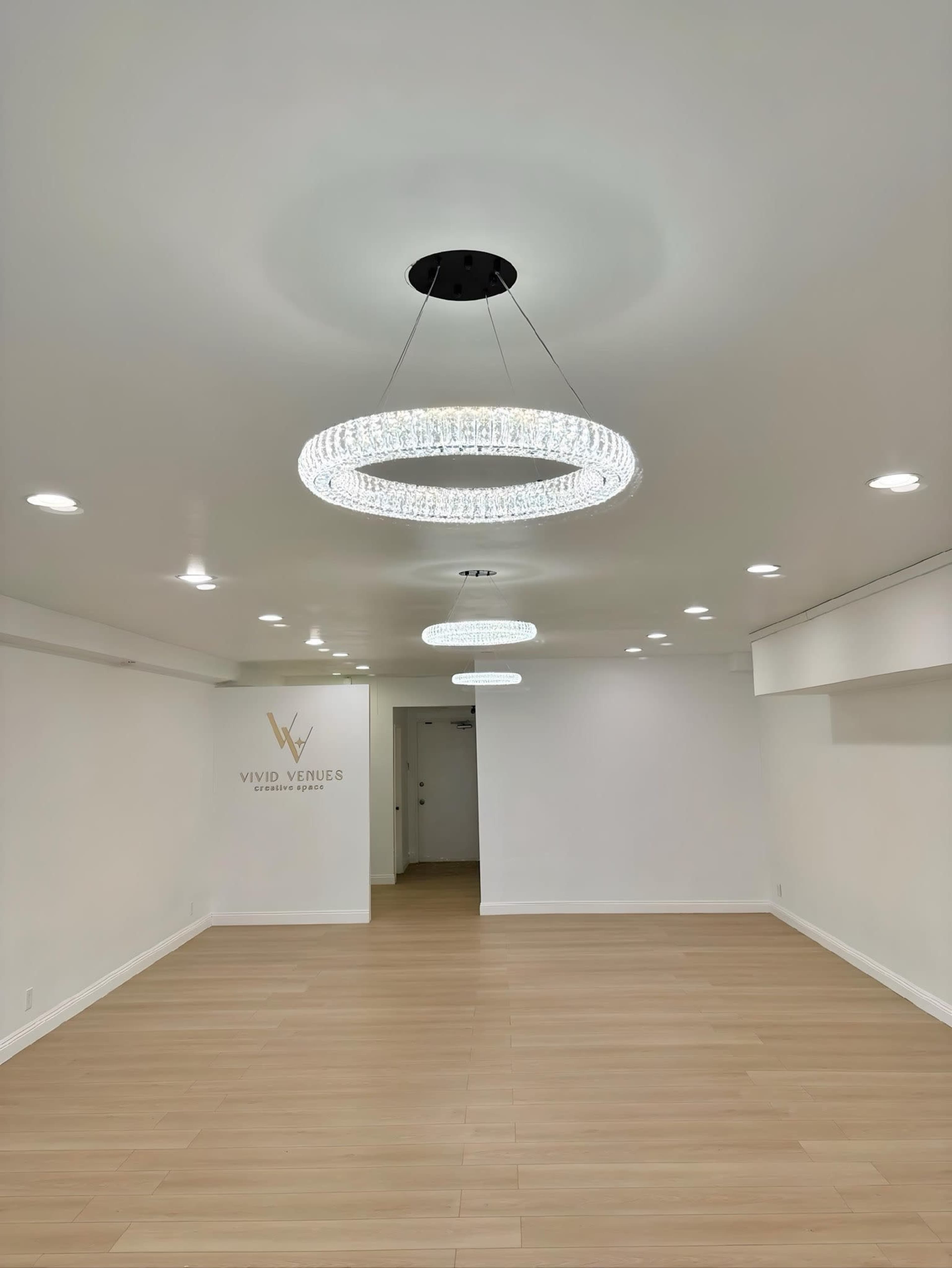 The image shows a spacious, empty room featuring two circular crystal chandeliers and a wall with the text "Vivid Venues."