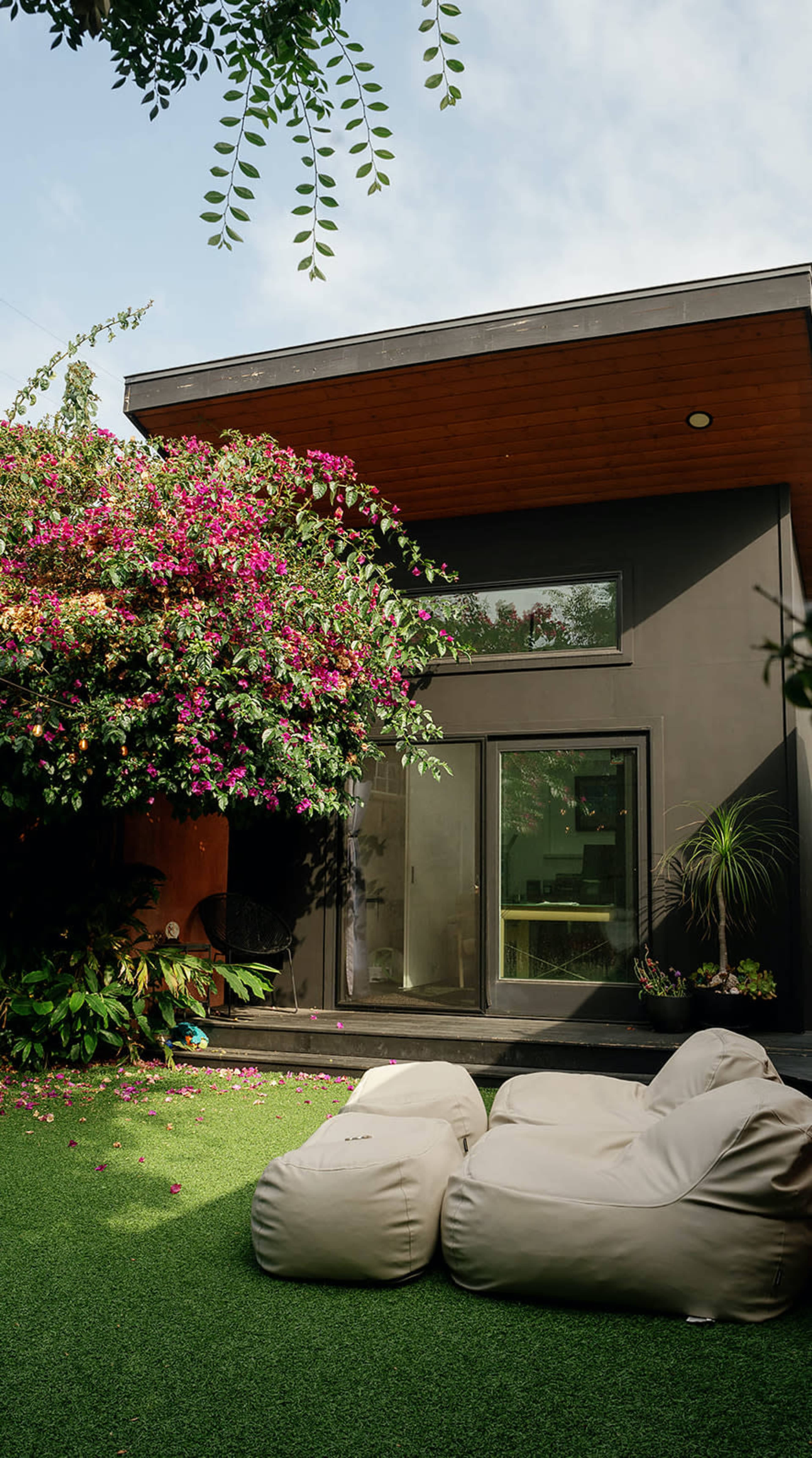 A modern home with large glass doors is surrounded by vibrant flowering plants and features two light-colored bean bags on a lush green lawn.