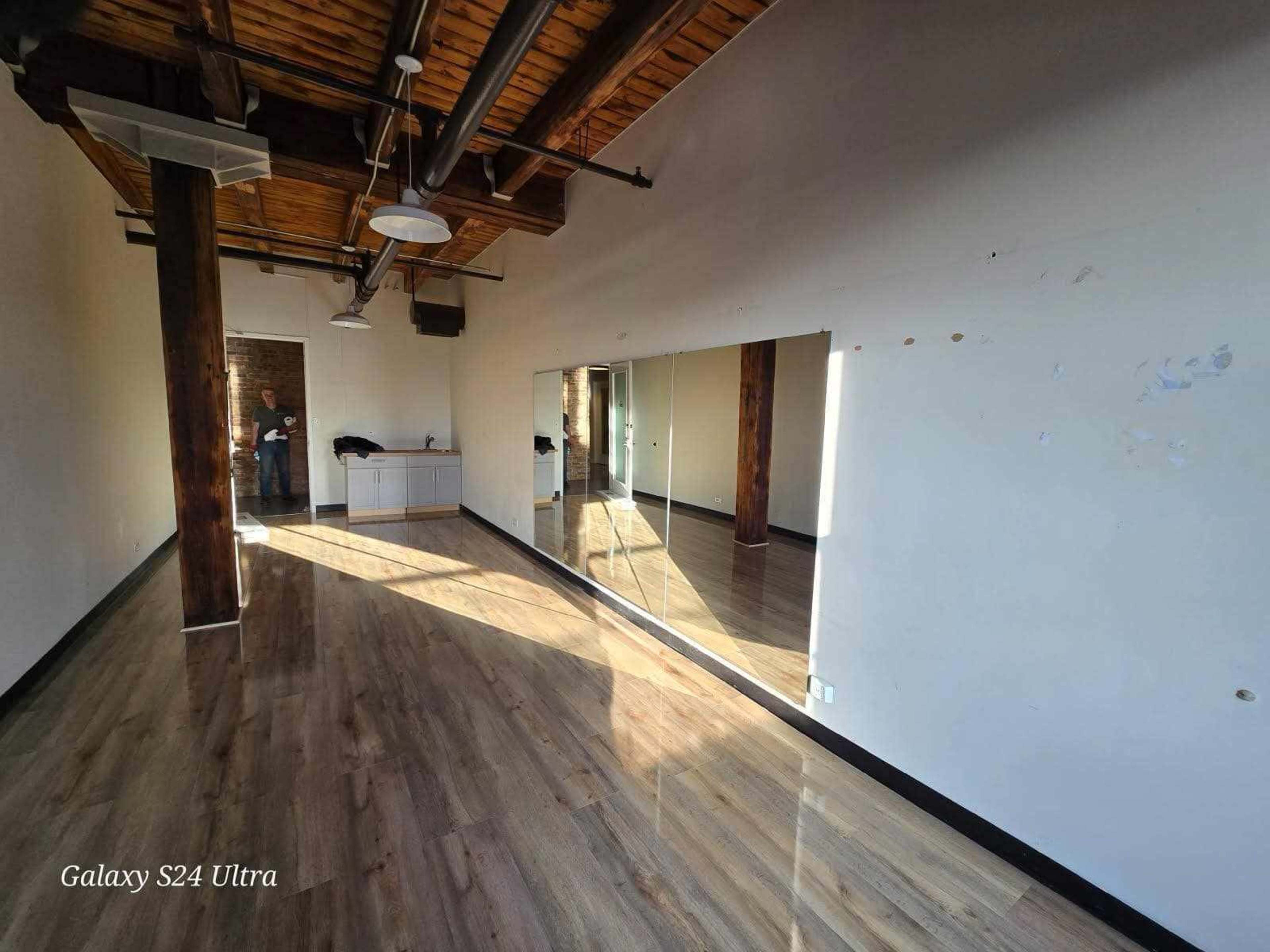 The image shows a spacious room with wooden beams and a large mirror on one wall, featuring polished flooring and minimal furnishings.