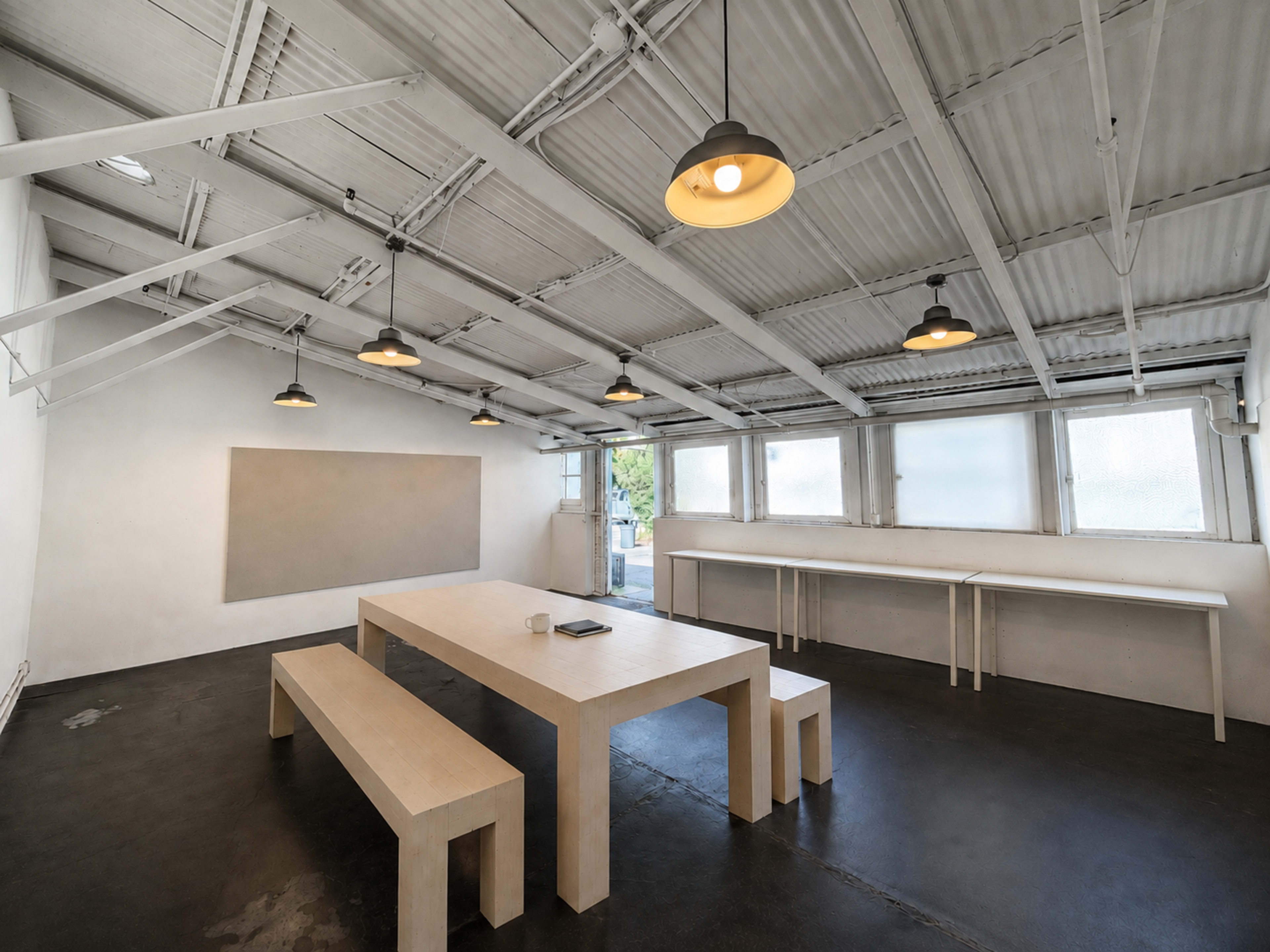 The image shows a minimalist meeting room with a large wooden table, several overhead lights, and large windows allowing natural light to enter.