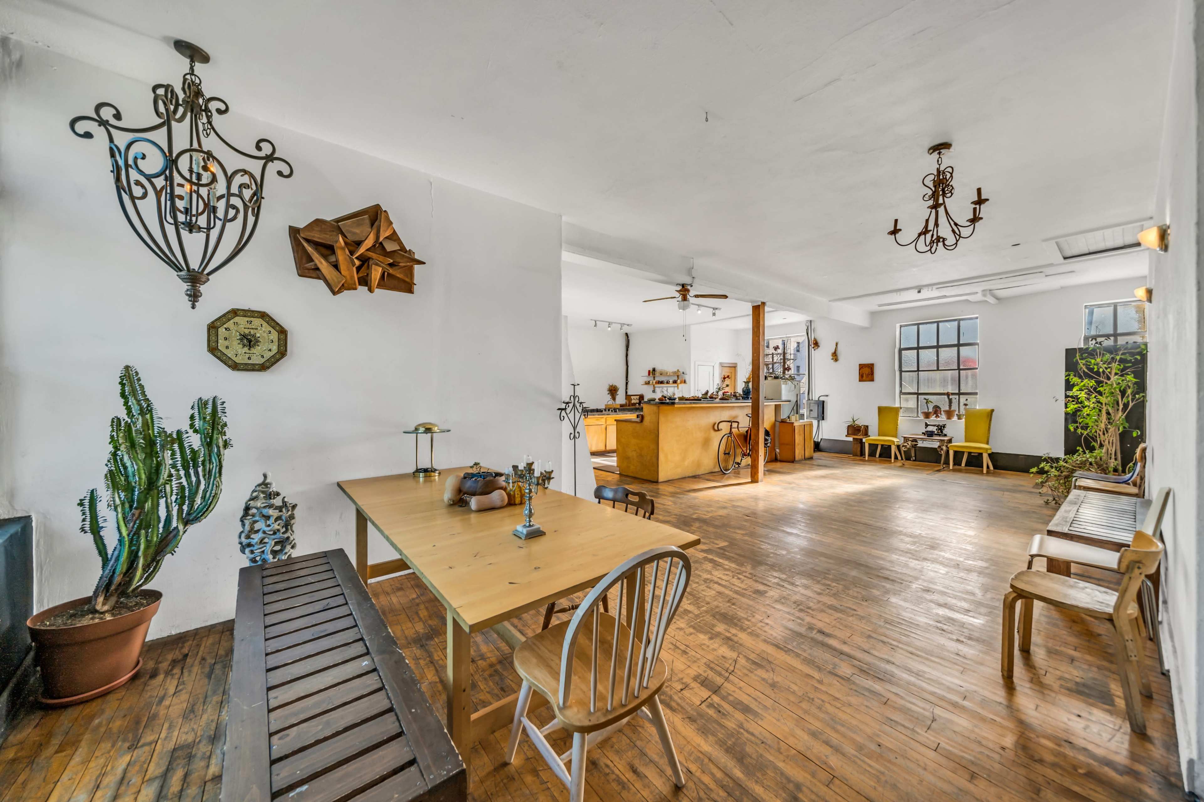 The image shows a spacious, airy room with wooden floors, a large central table, and a bar area, adorned with various decorations and seating arrangements.
