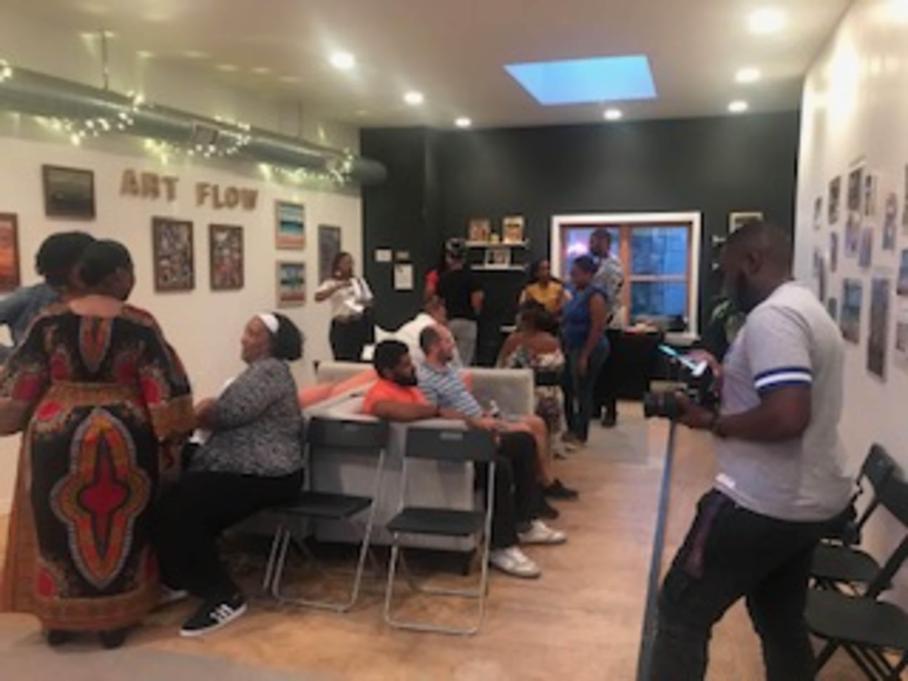 A group of people is gathered in an art space, engaging in conversation and taking photos, with artwork displayed on the walls.