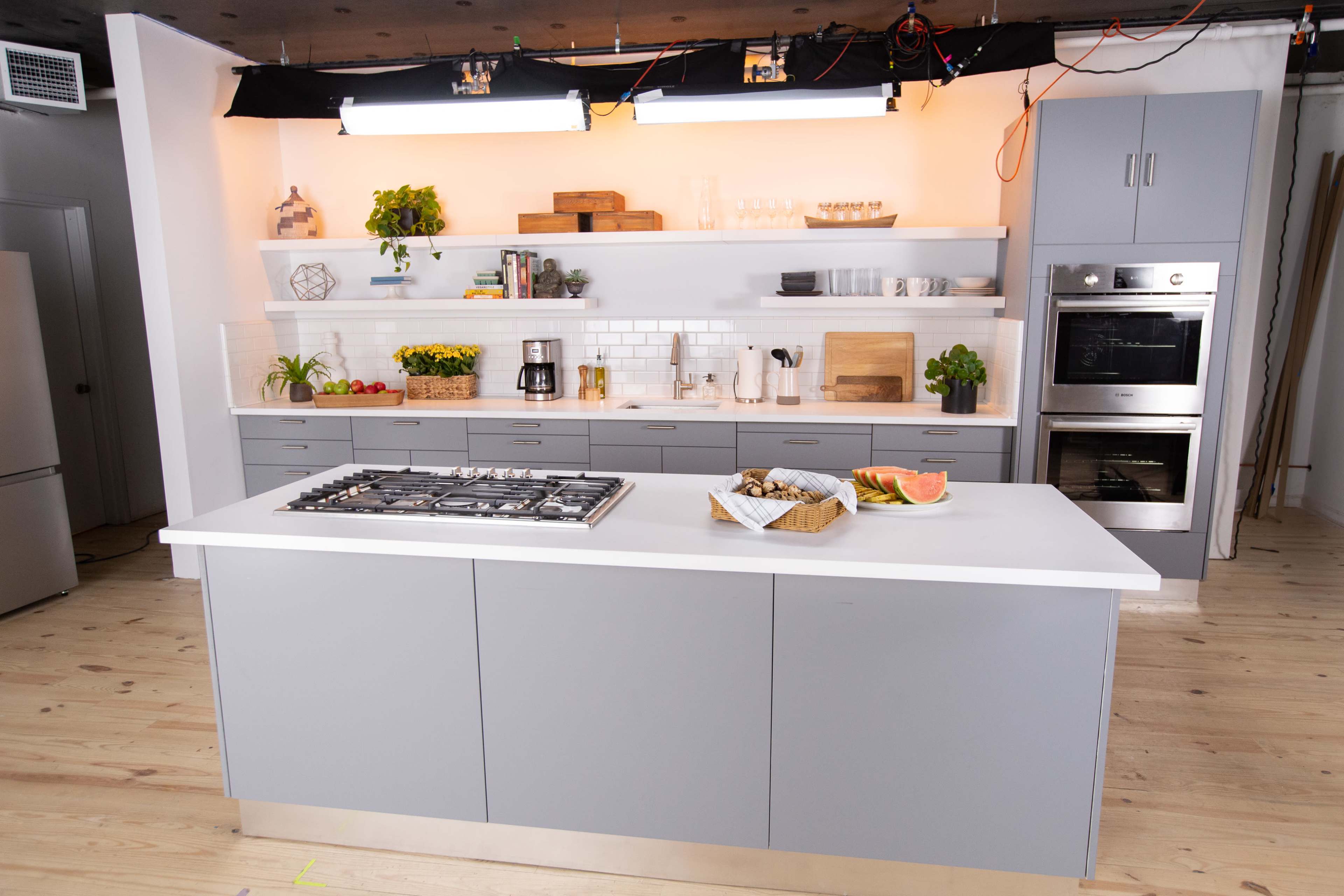 A modern kitchen features a central island with a gas cooktop, surrounded by minimalist cabinetry and shelves displaying various kitchen items.