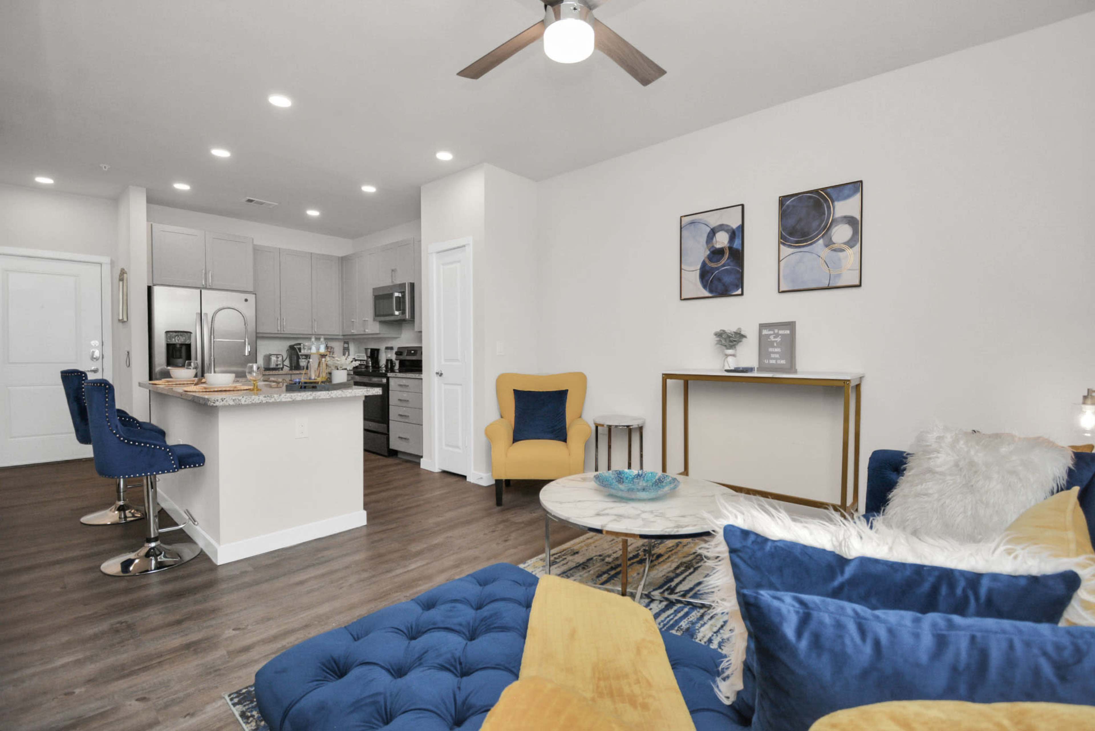 The image shows a modern living room with a blue and yellow color scheme, featuring a sofa, a coffee table, and a kitchen in the background.