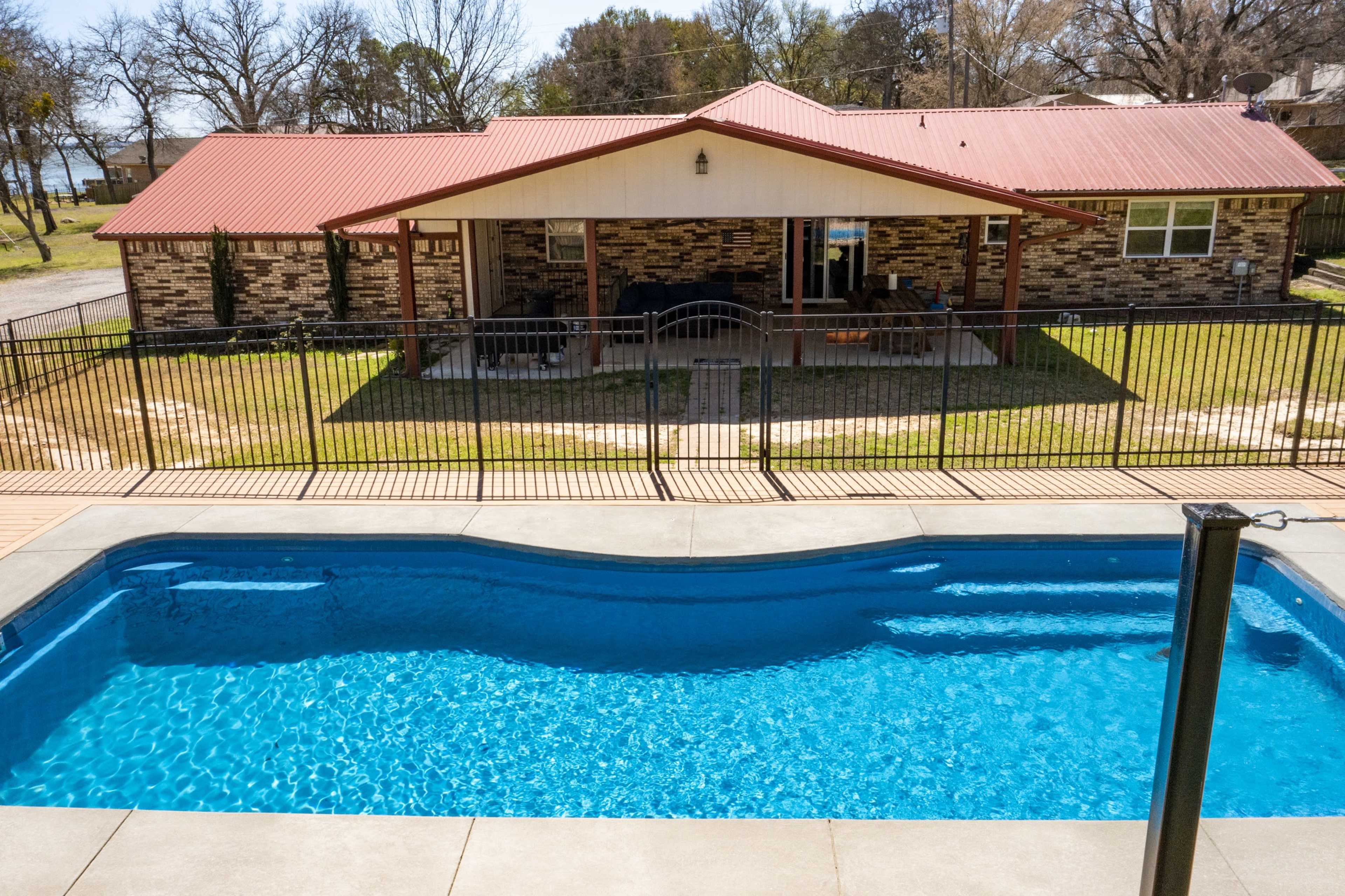 A backyard featuring a swimming pool in the foreground and a house with a red roof and stone walls in the background, surrounded by a fence.