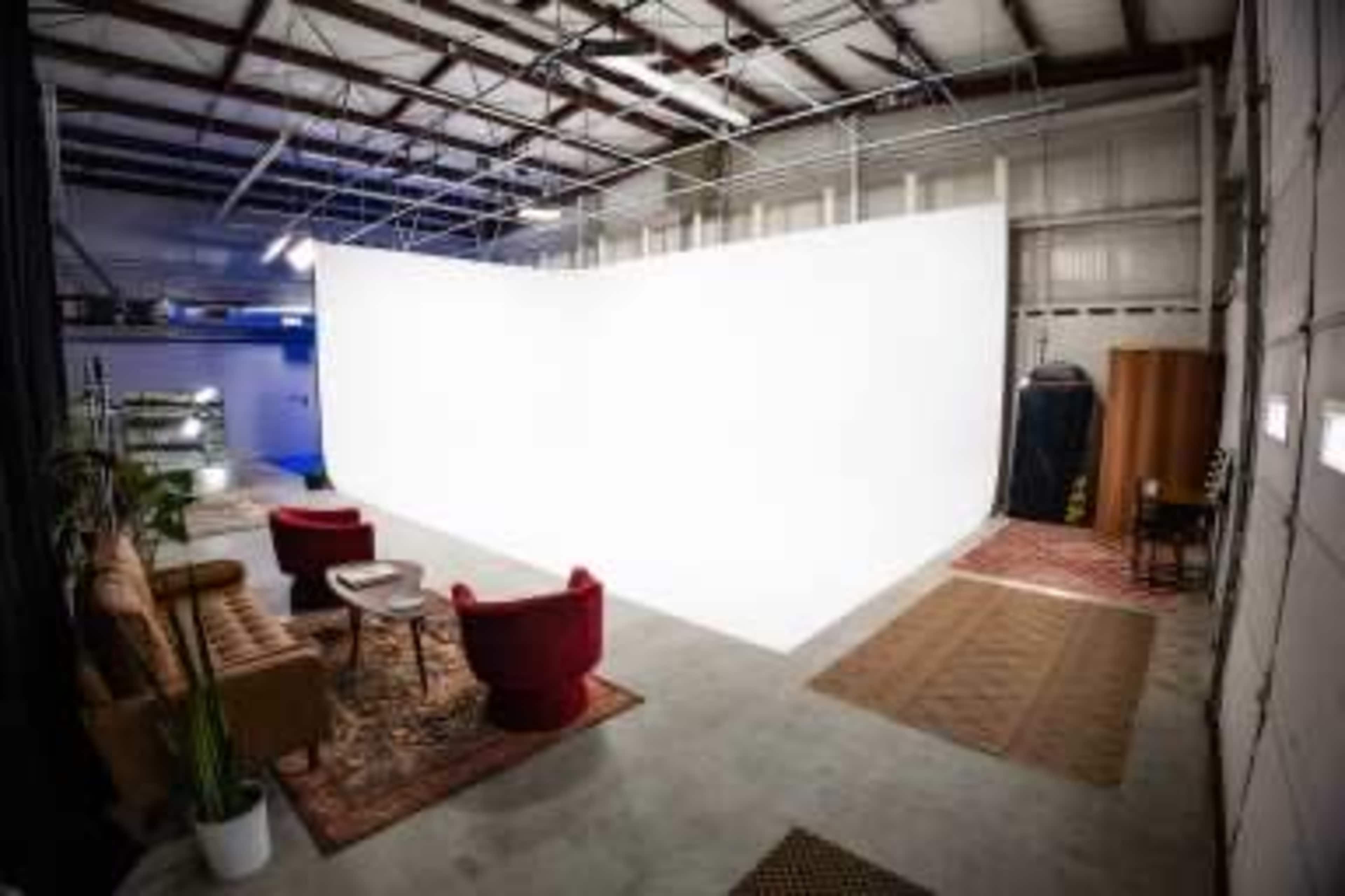 The image shows an indoor studio space featuring a white backdrop, a sofa, two red chairs, and decorative rugs on the concrete floor.