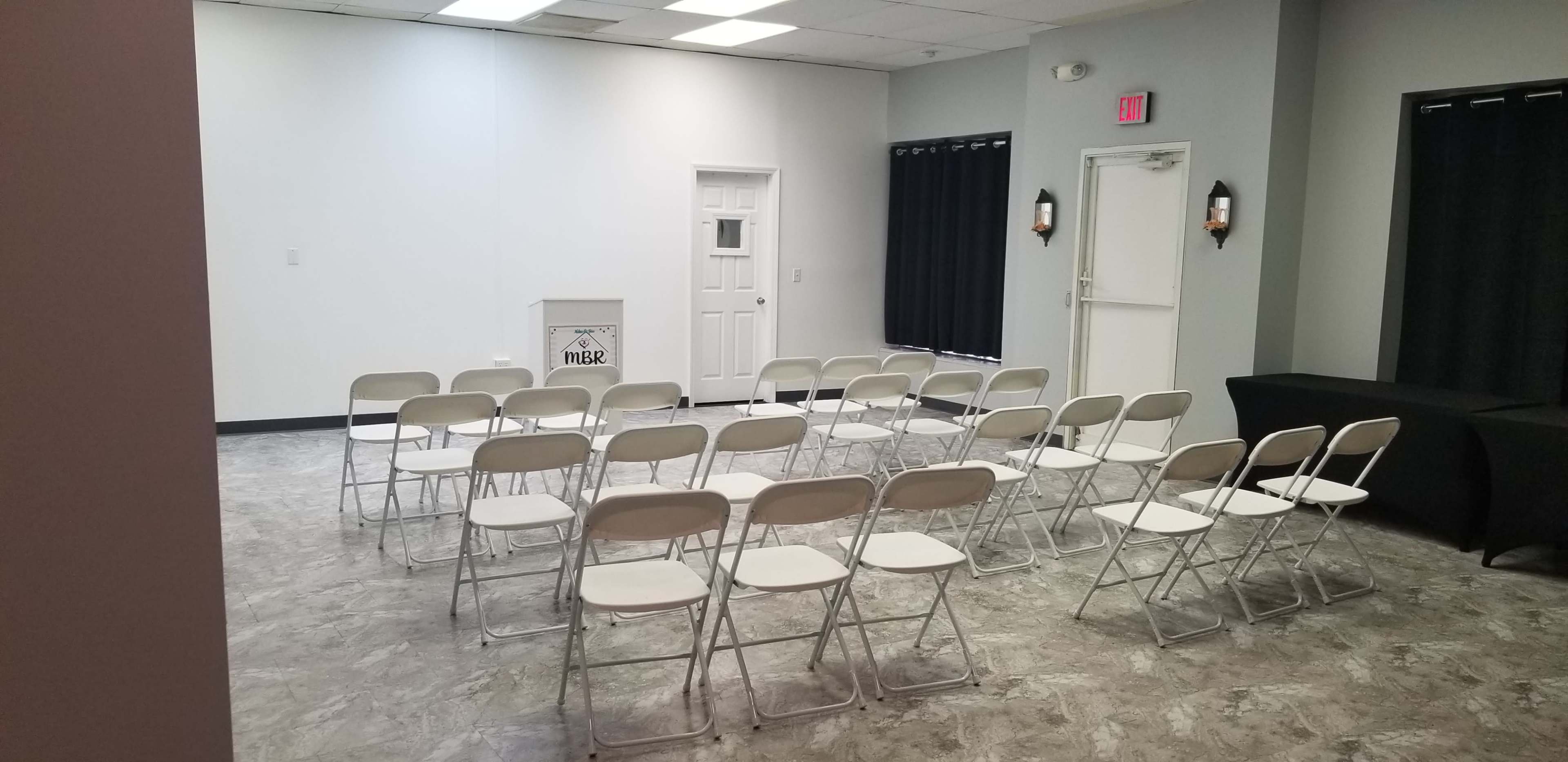 The image shows a room with a row of white folding chairs set up facing a blank wall, with a door and dark curtains on the sides.