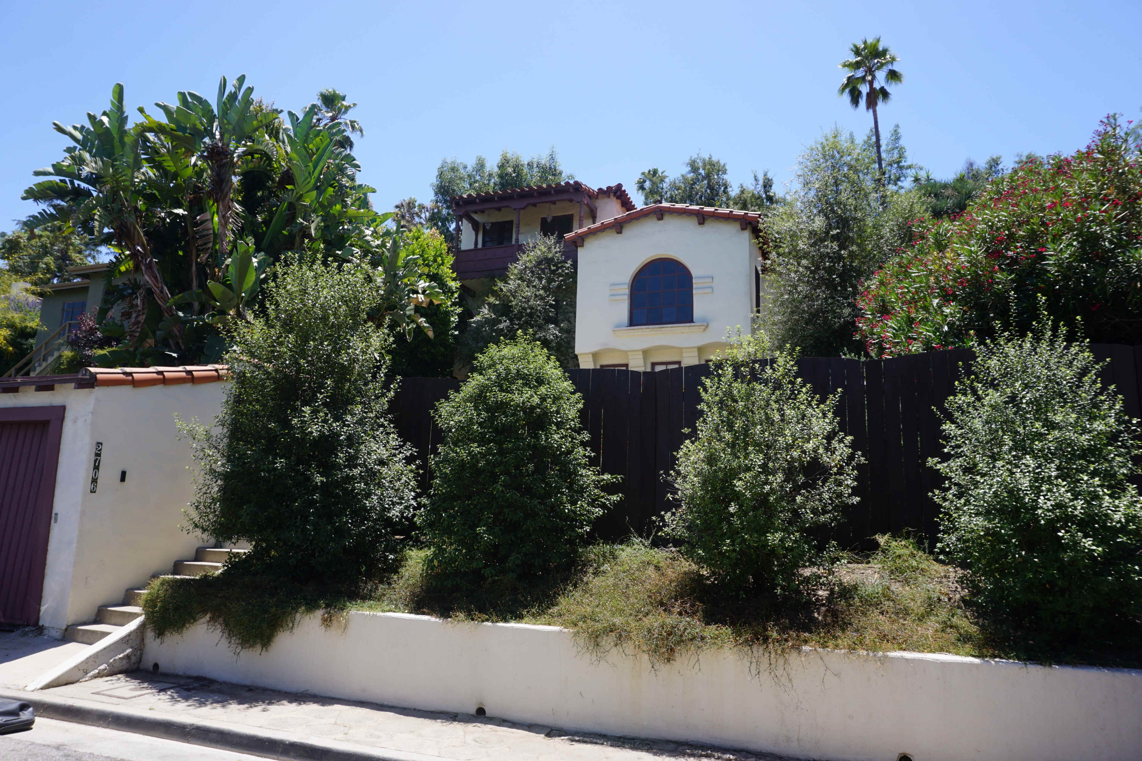 A hillside home is surrounded by lush greenery and shrubs, with a wooden fence in the background.