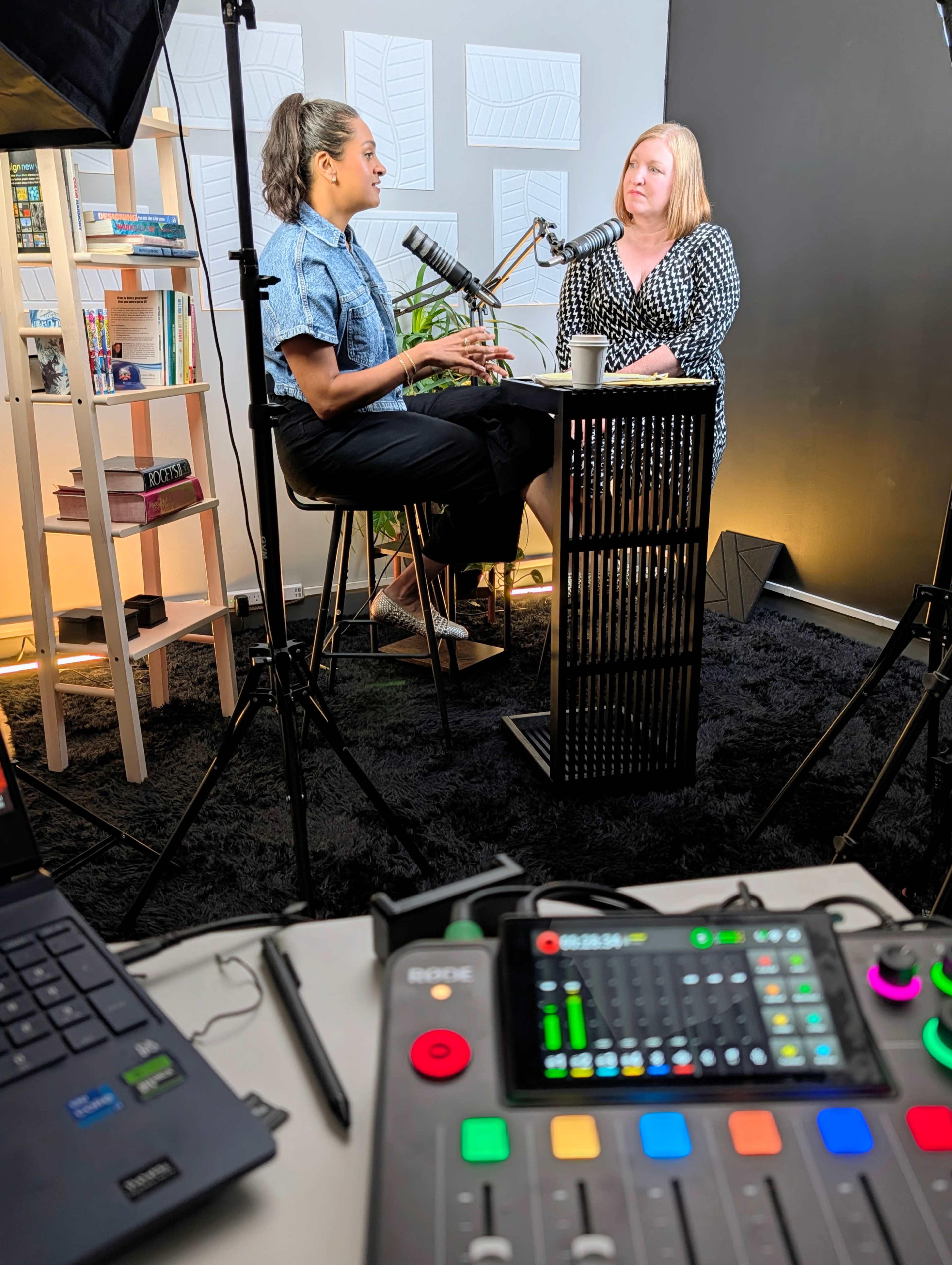 Two women are seated at a podcast recording setup, speaking into microphones in a well-lit studio.