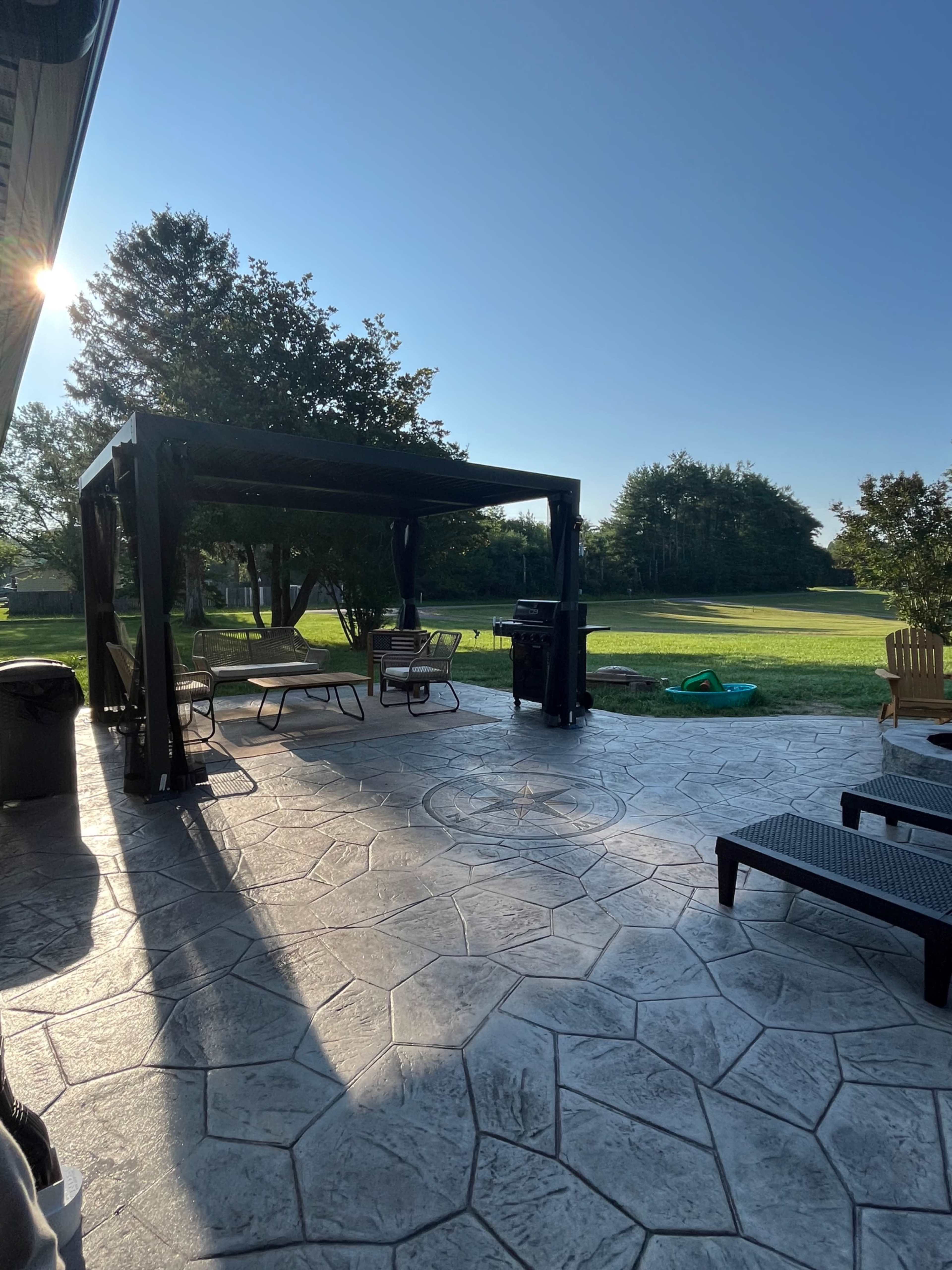 A patio area features a black pergola, outdoor furniture, and a grill, set against a backdrop of a grassy yard and trees under a clear blue sky.