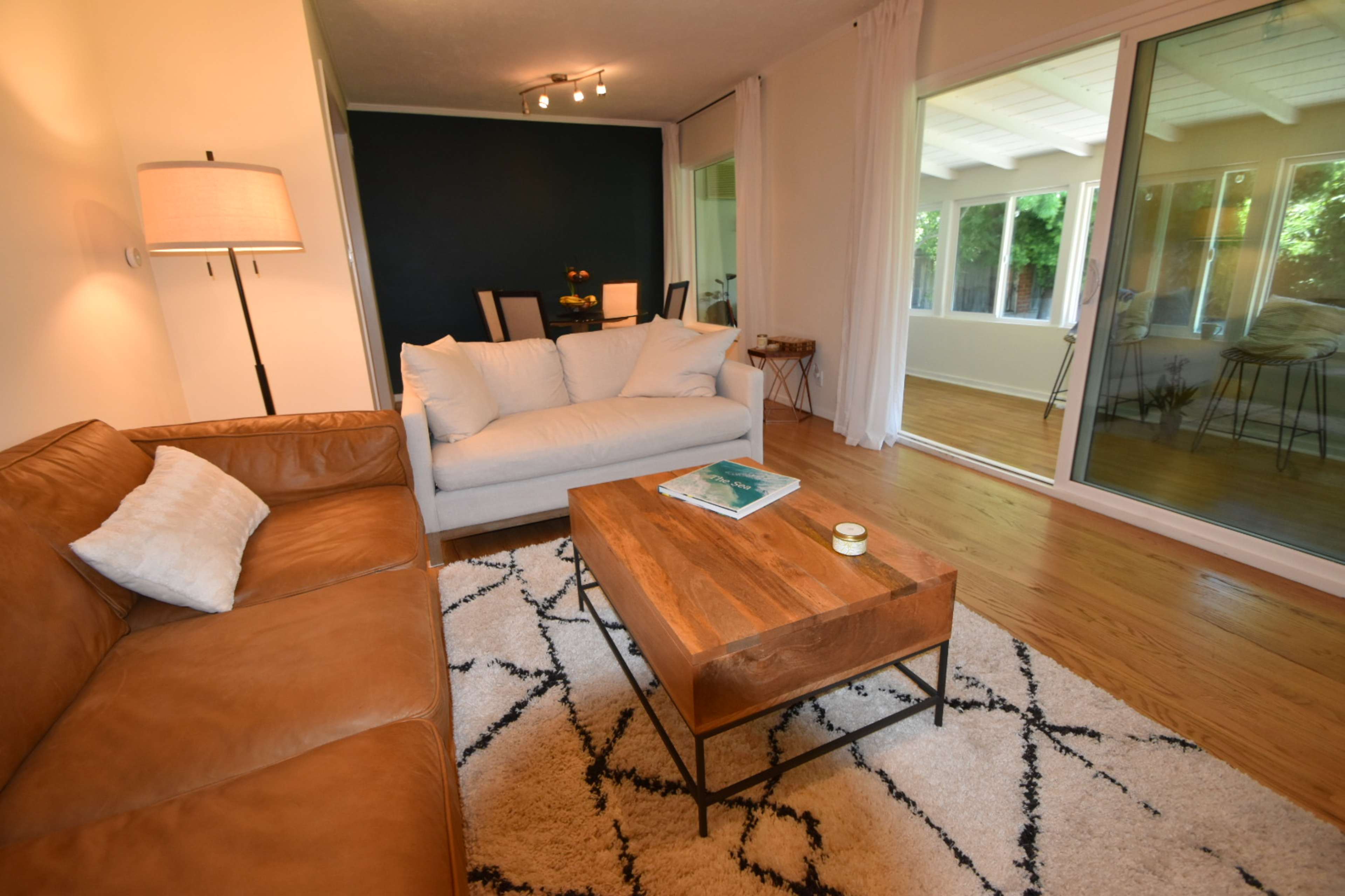 The image shows a living room with a tan leather sofa, a light-colored sectional sofa, a wooden coffee table, and large windows that provide natural light.