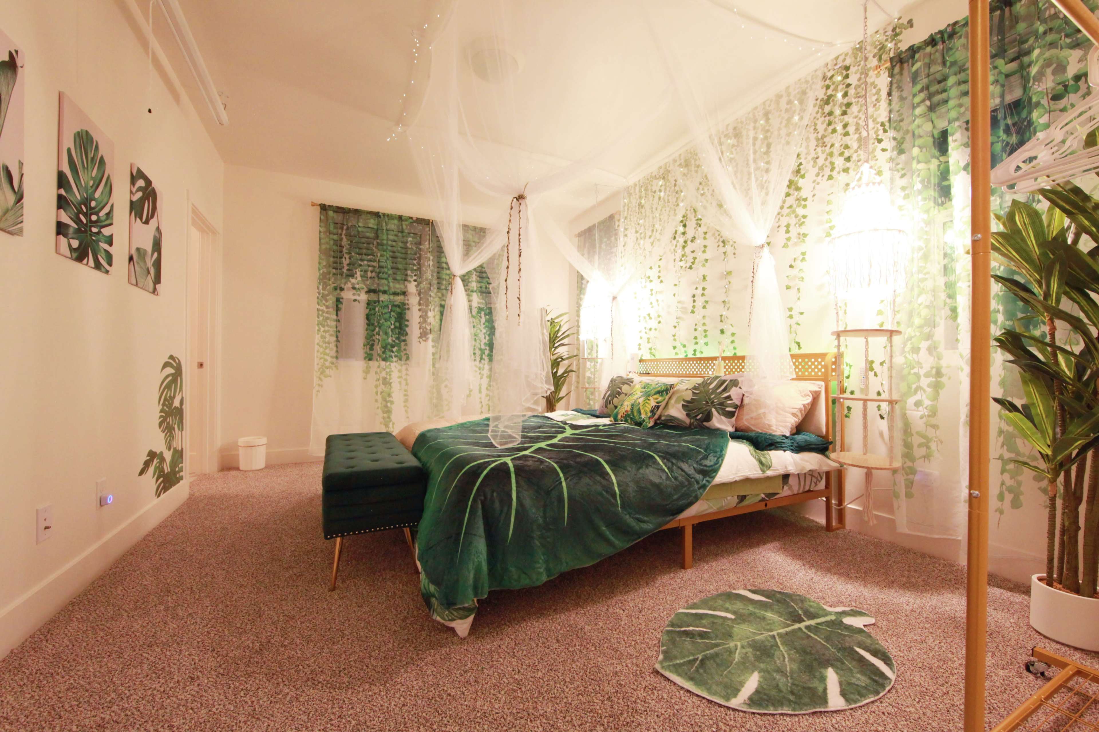 The image shows a bedroom decorated with green tropical-themed bedding and wall art, featuring plants and sheer curtains.