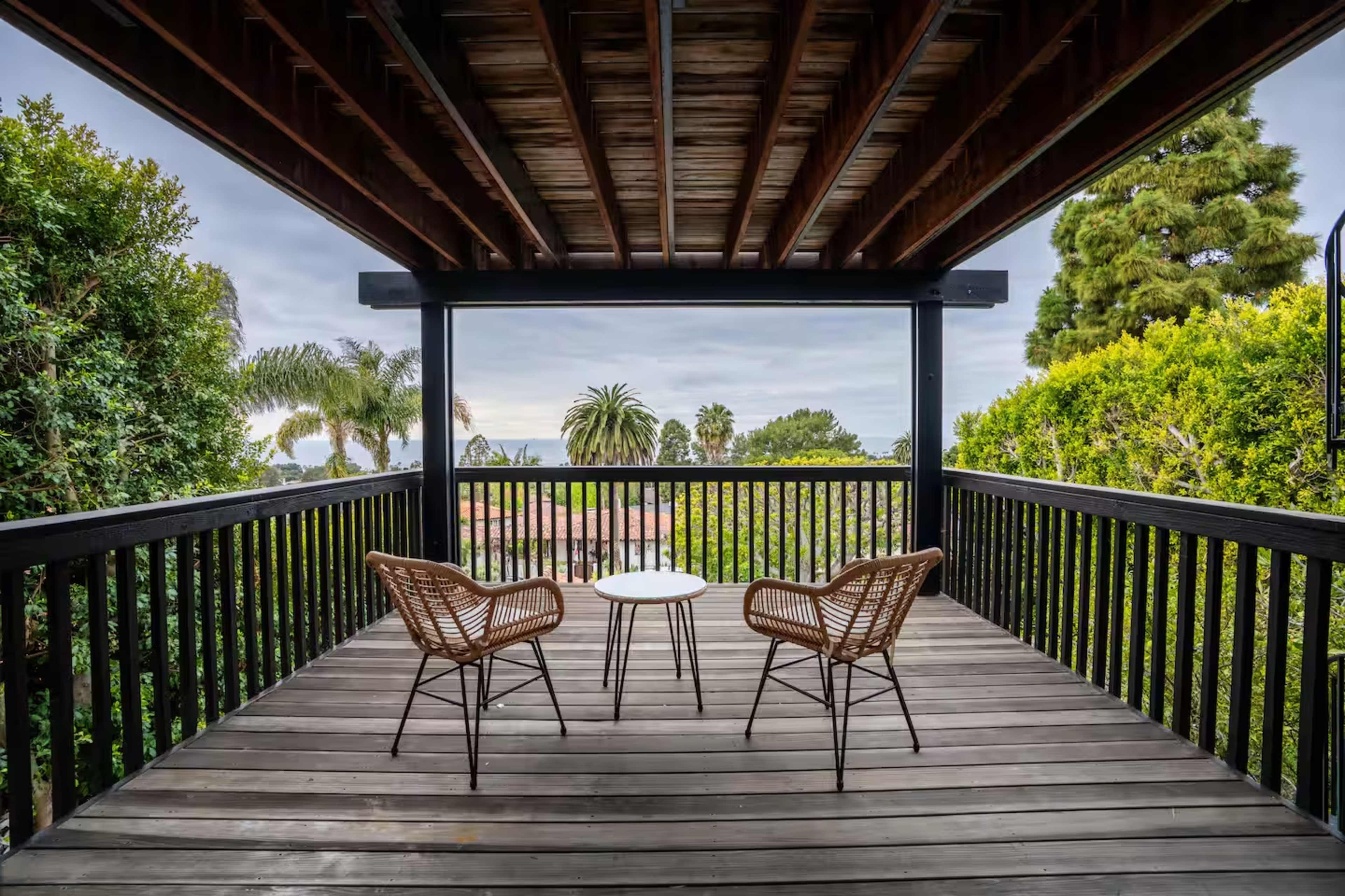 A wooden deck features two woven chairs and a round table, surrounded by lush greenery and palm trees.