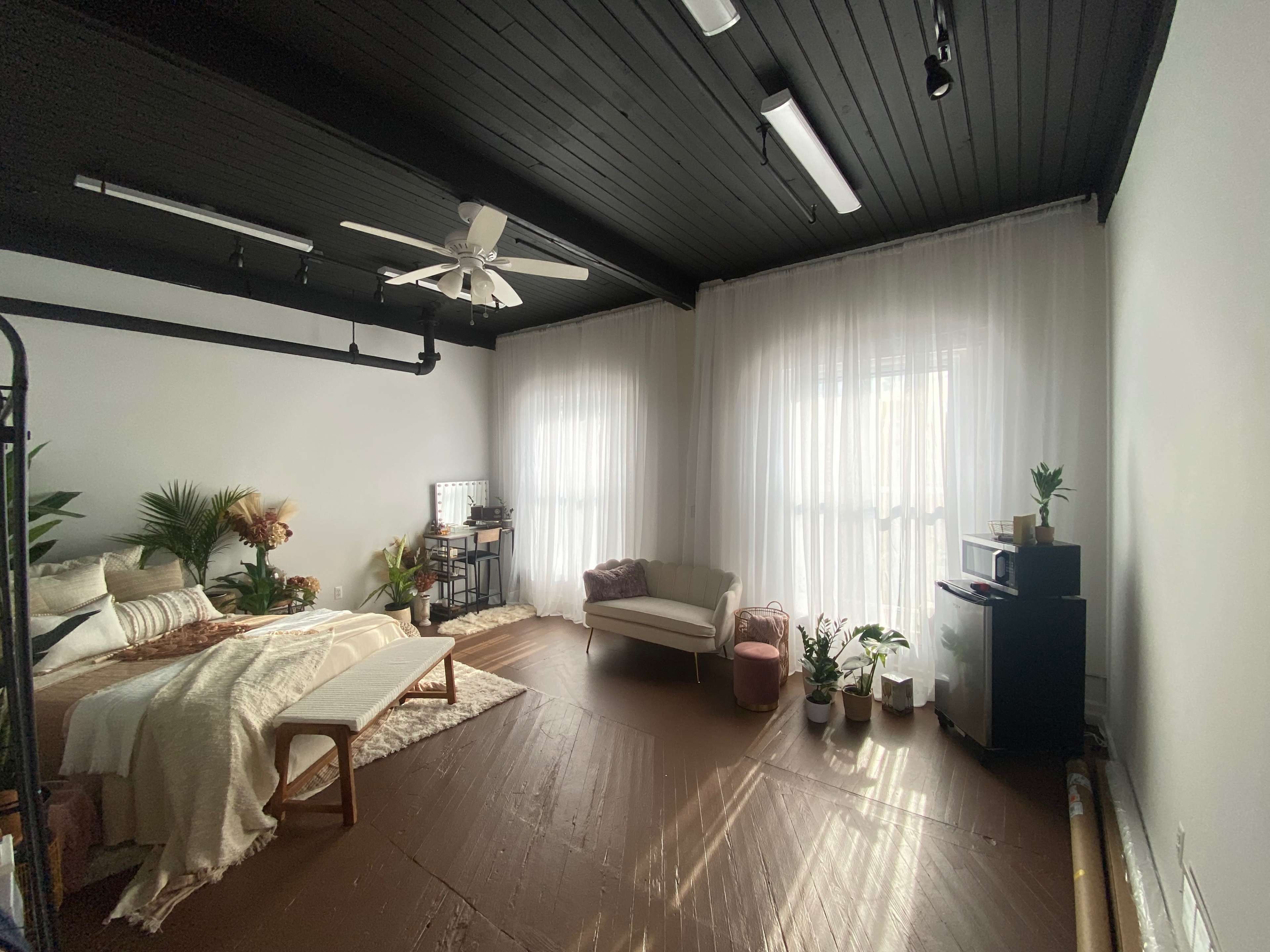 The image shows a spacious bedroom featuring a bed with light bedding, a couch, and several potted plants, illuminated by natural light through sheer curtains.