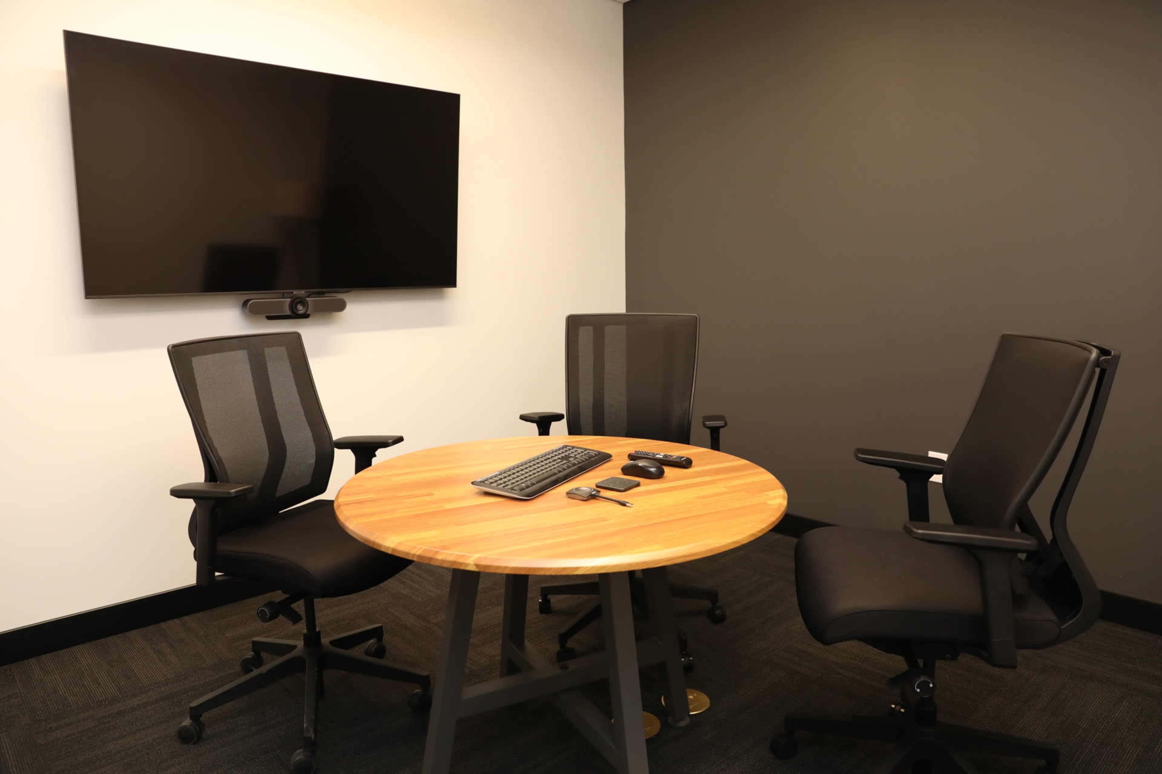 A small meeting room features a circular wooden table with three black chairs and a large wall-mounted screen.