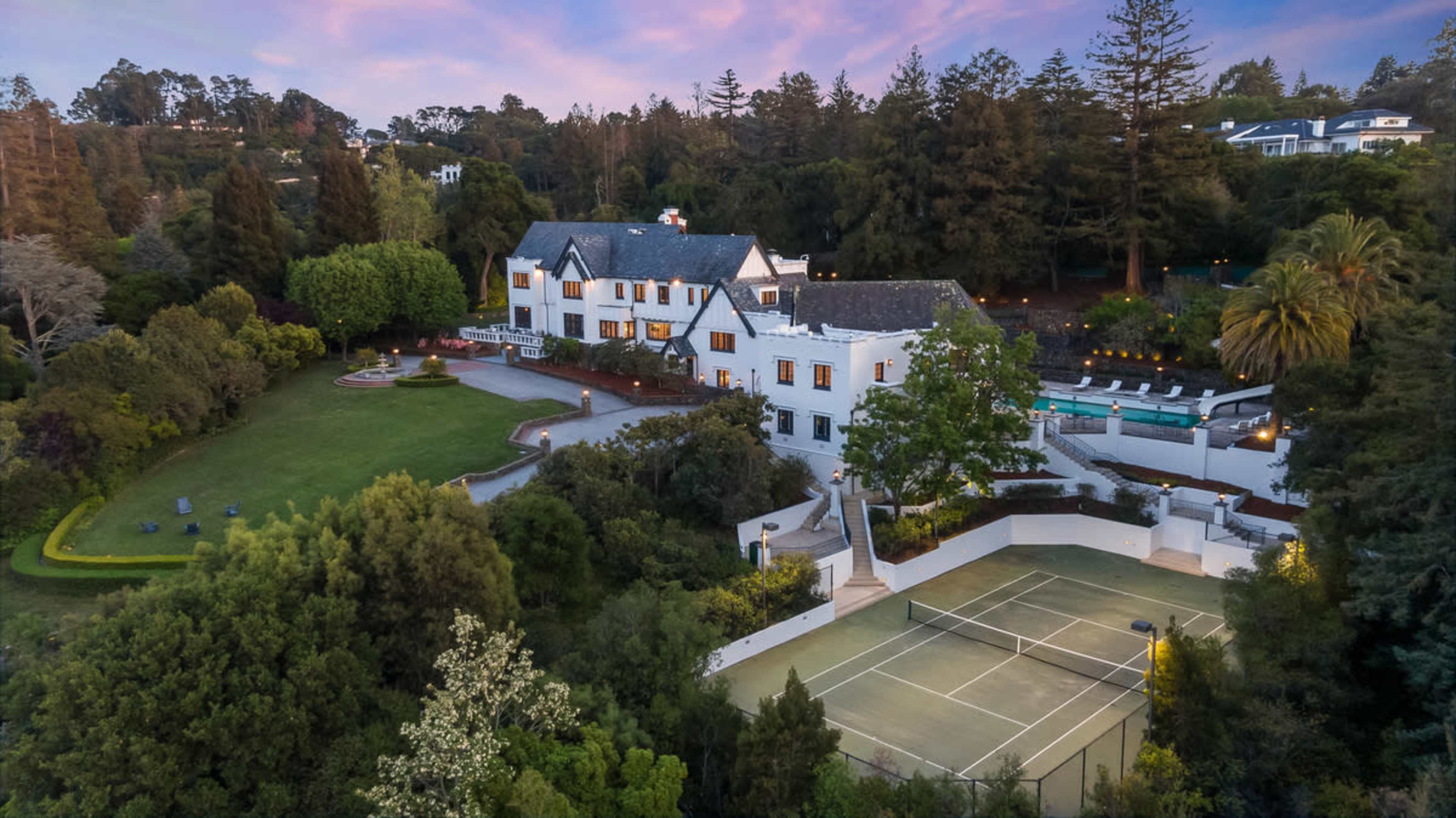 A large white mansion is set amidst lush greenery, with a swimming pool and a tennis court adjacent to the property.