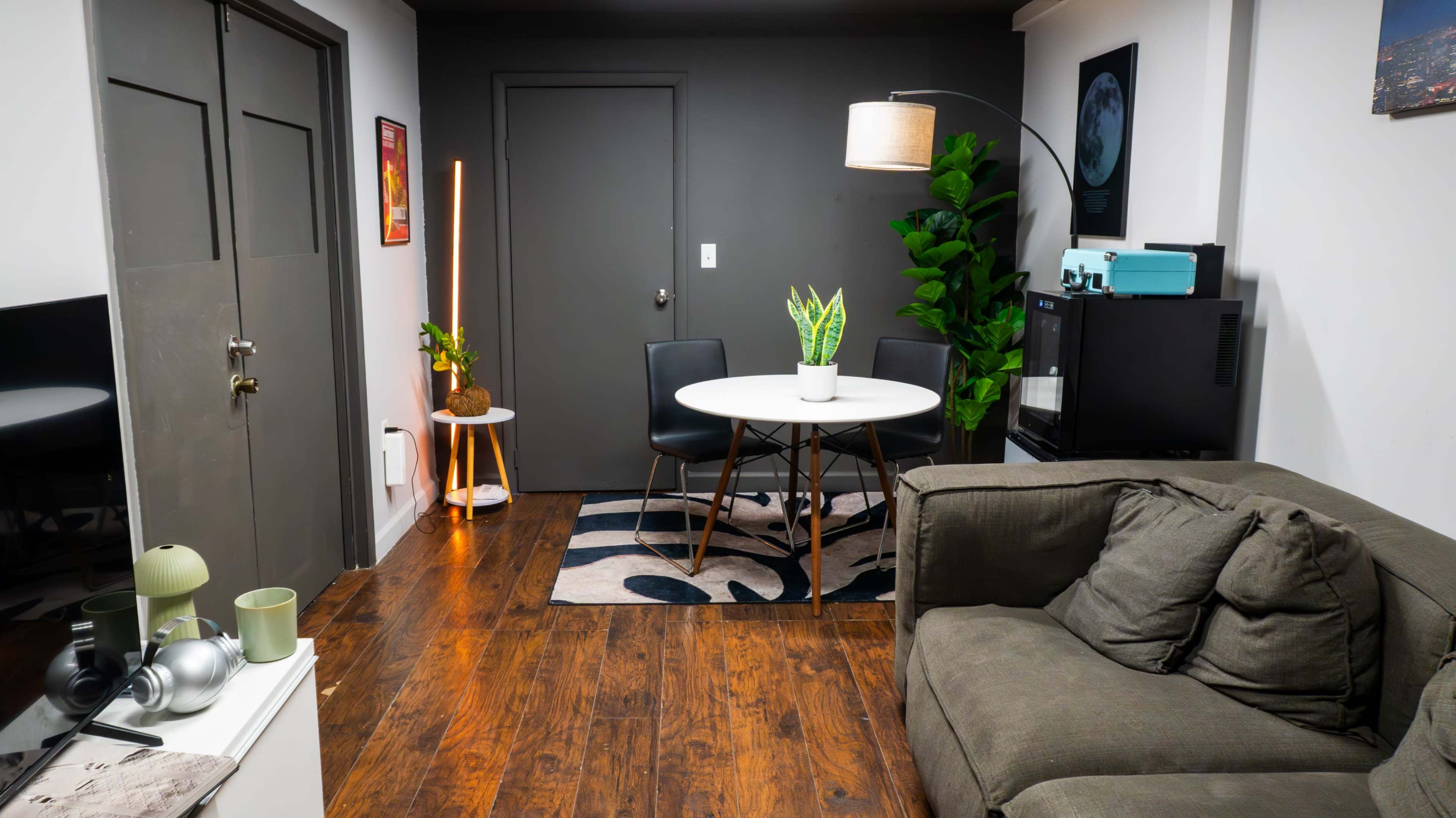 A small living space features a round white dining table with black chairs, a green potted plant, and a couch, alongside a wooden floor and modern decor.