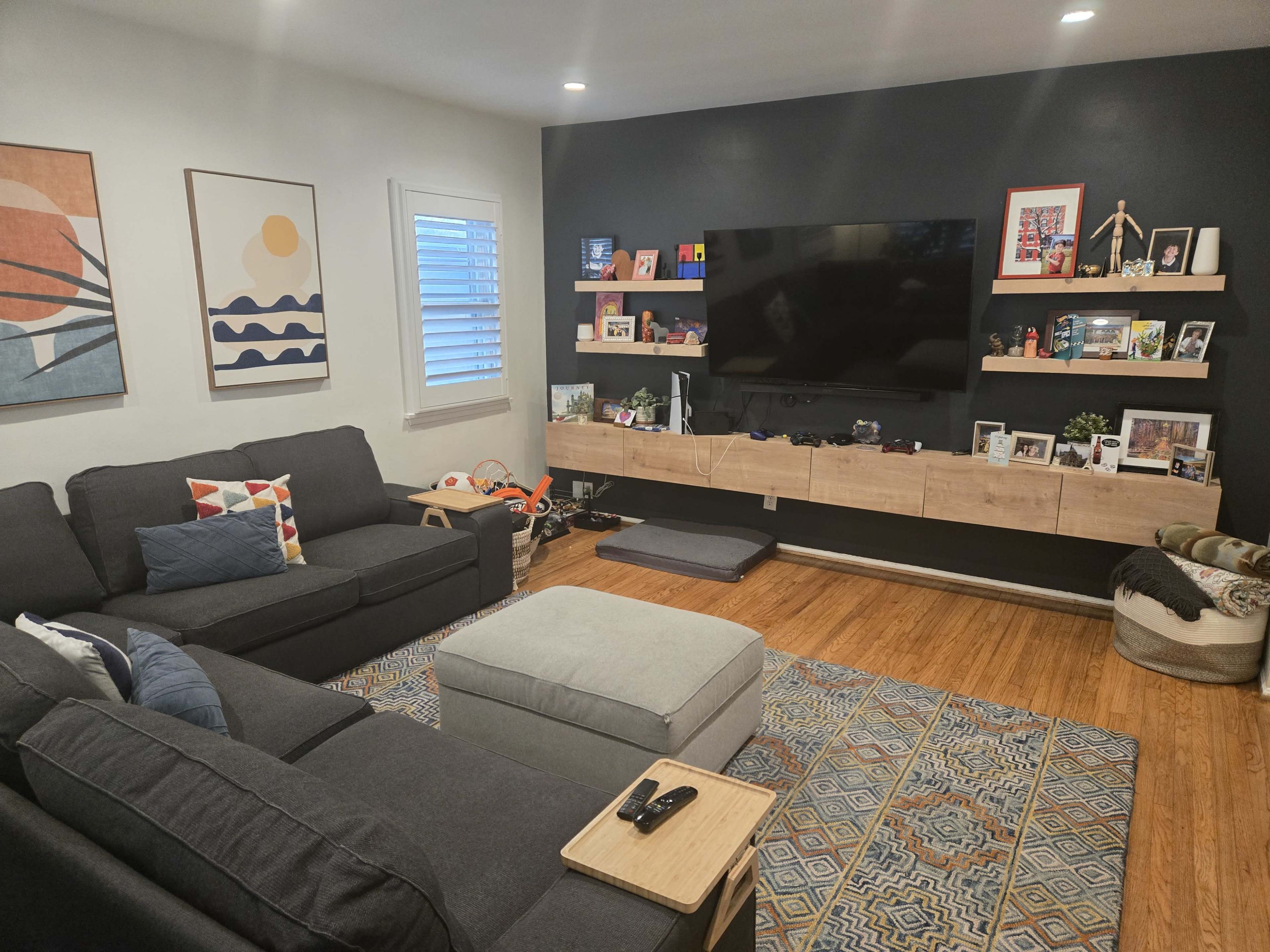 The living room features two dark couches, a light gray ottoman, a wall-mounted television, and decorative shelves displaying photos and small items.