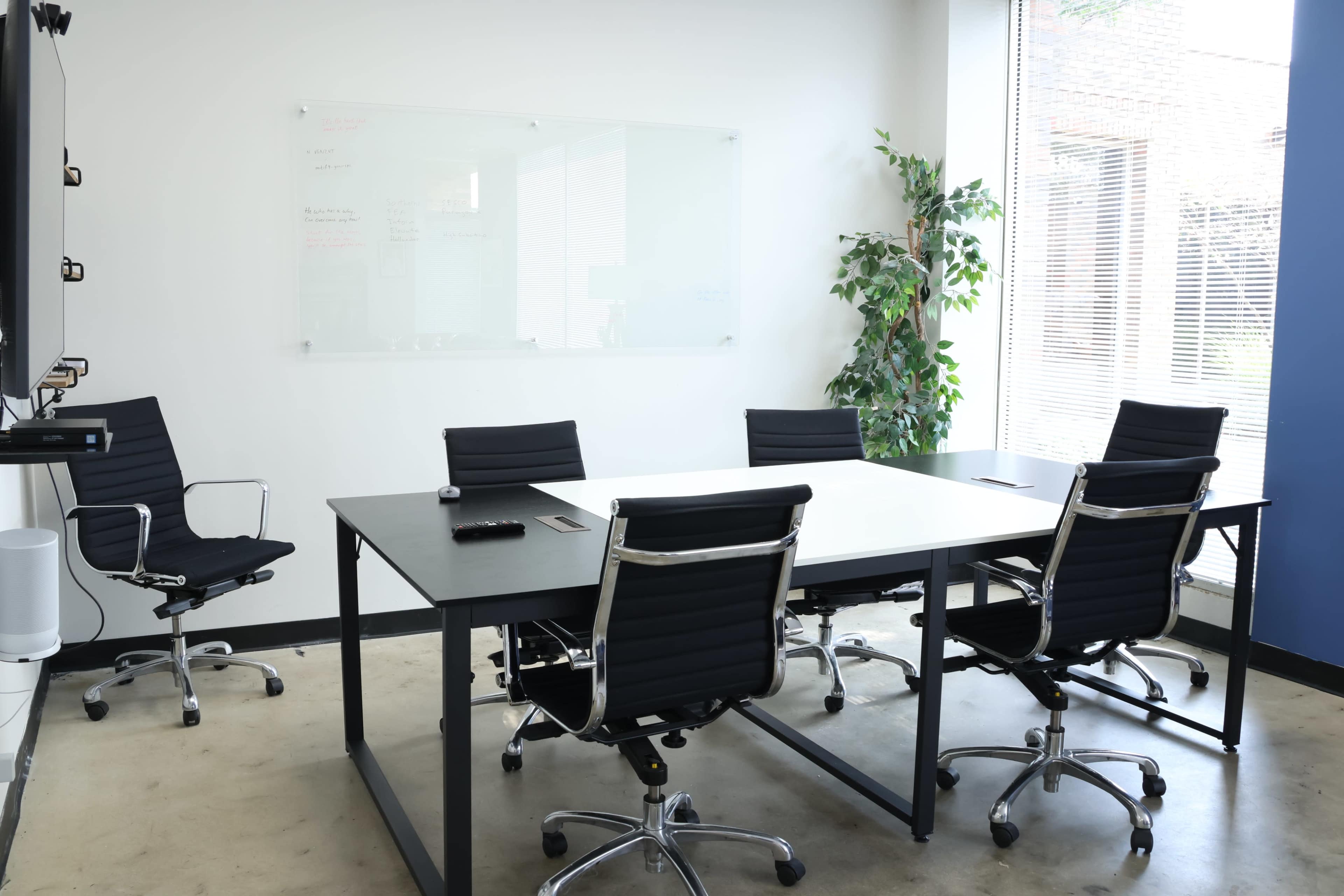 A modern conference room features a large table surrounded by five ergonomic chairs, with a whiteboard and a potted plant in the background.