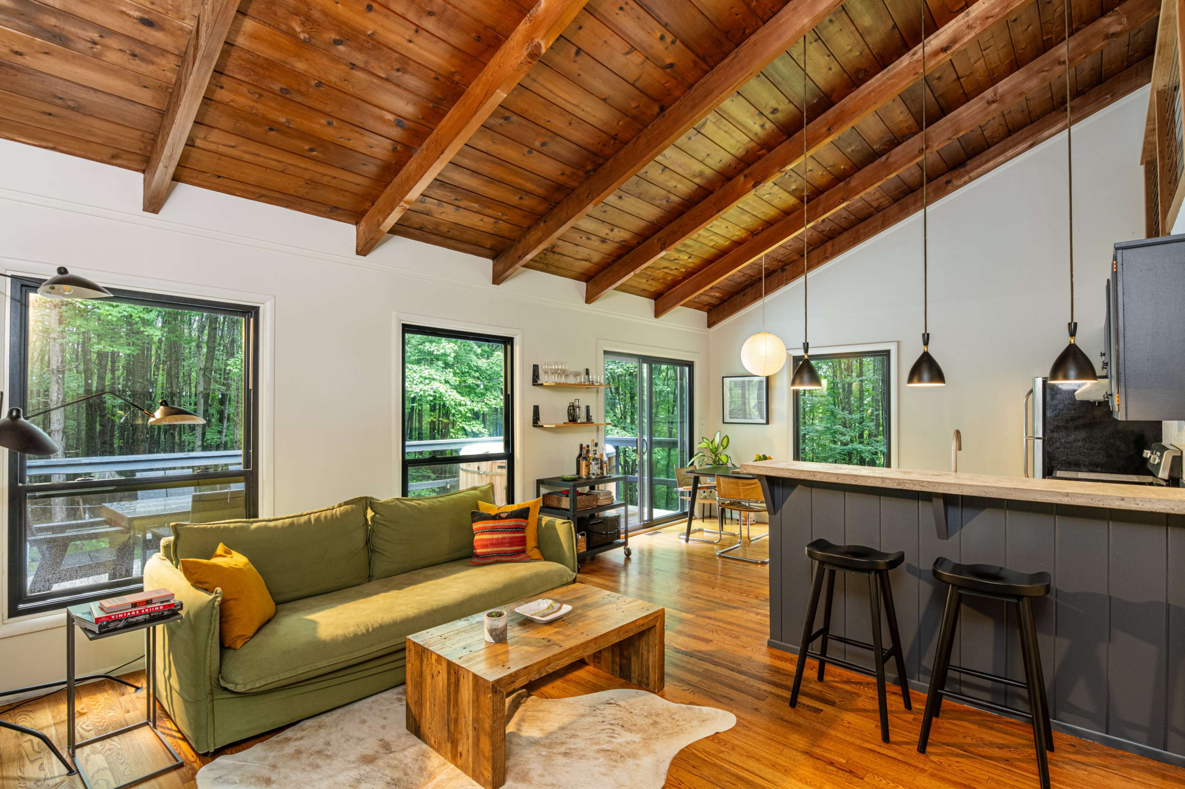 The interior of a modern living space features a green sofa, a wooden coffee table, and a kitchen area with a bar, all set against a backdrop of large windows overlooking a forest.