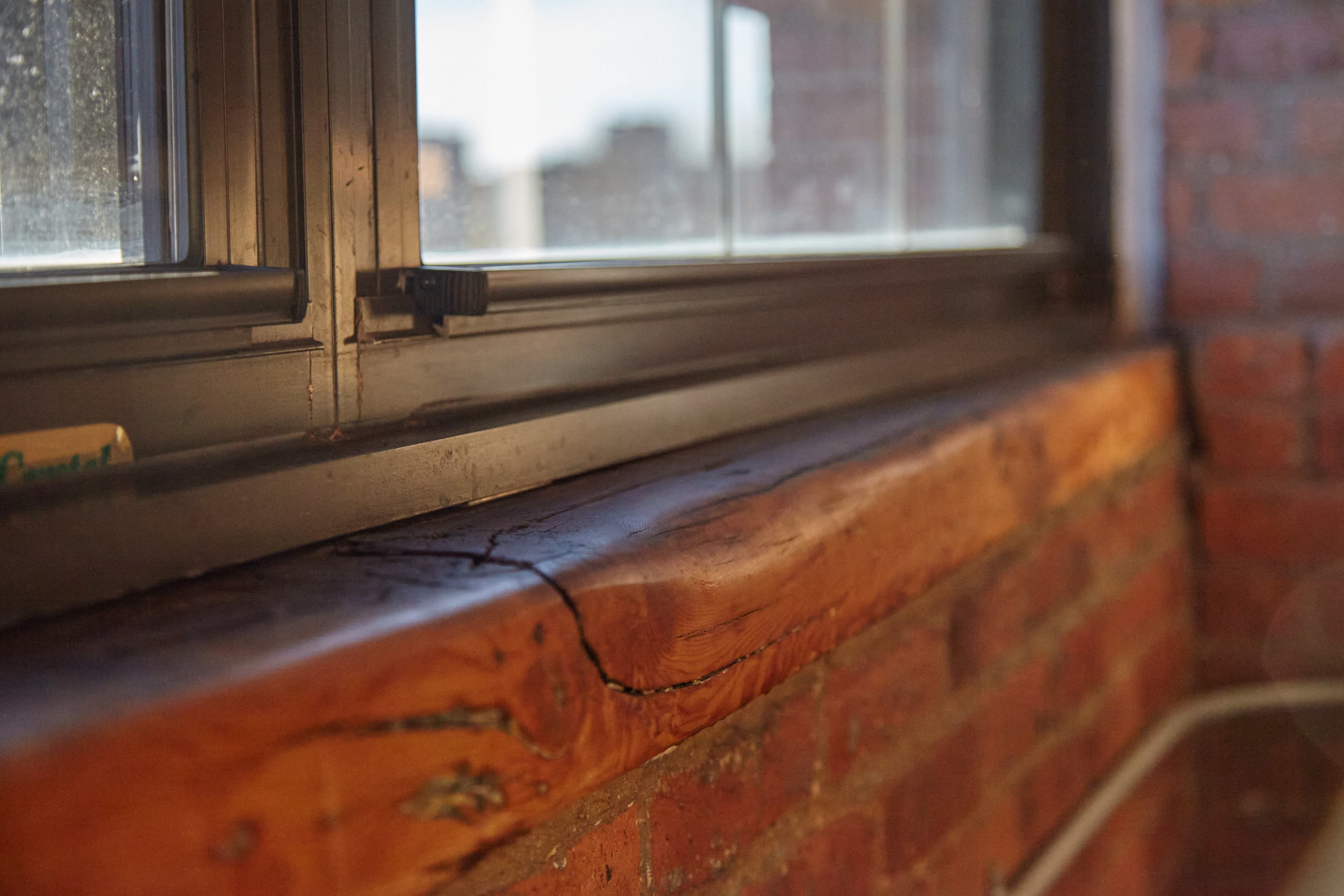 A wooden windowsill, cracked and resting against a brick wall, with a window partially open behind it.