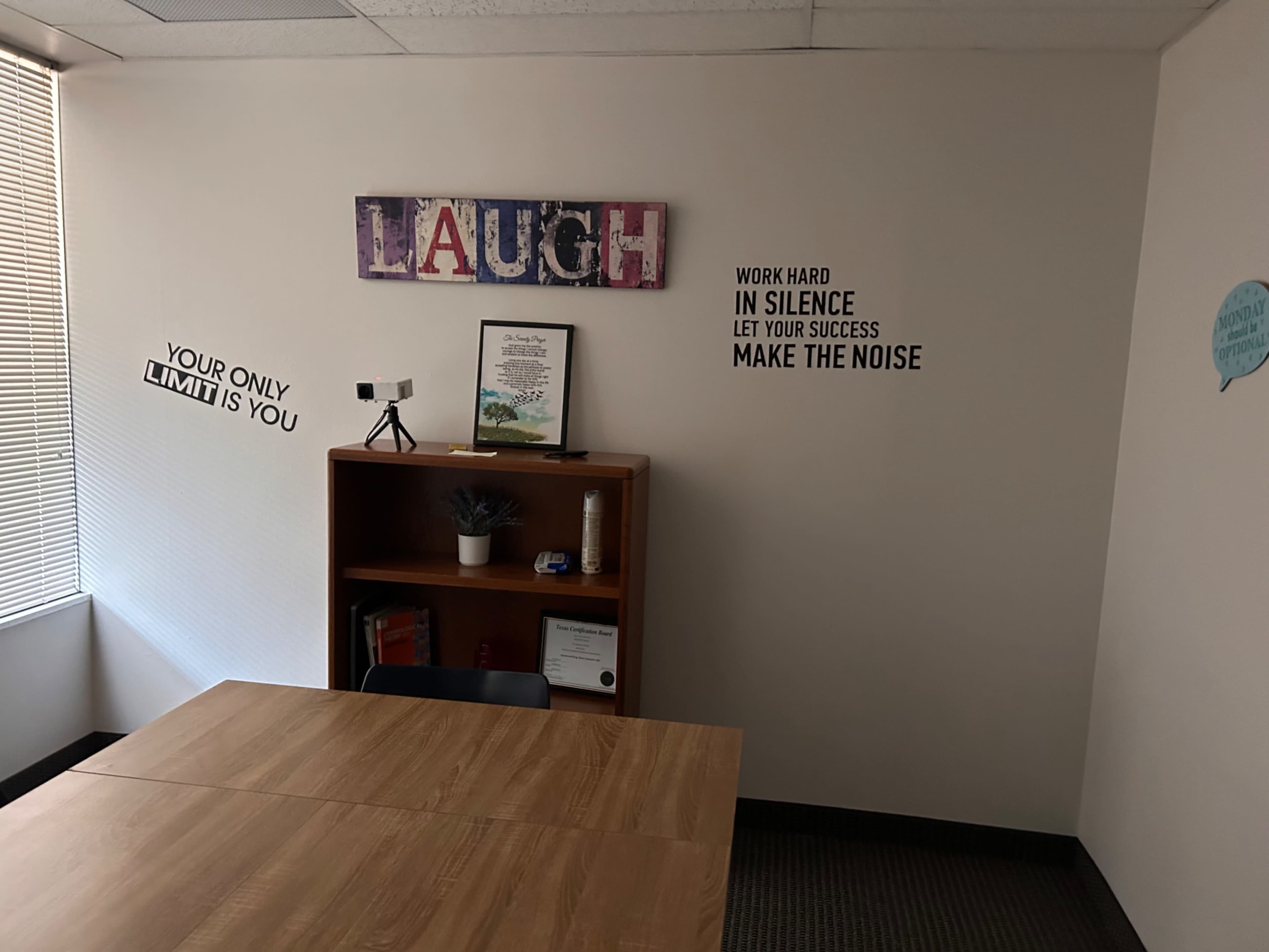 The image shows an interior office space with a wooden table, a bookshelf, motivational wall decor, and a window with blinds.