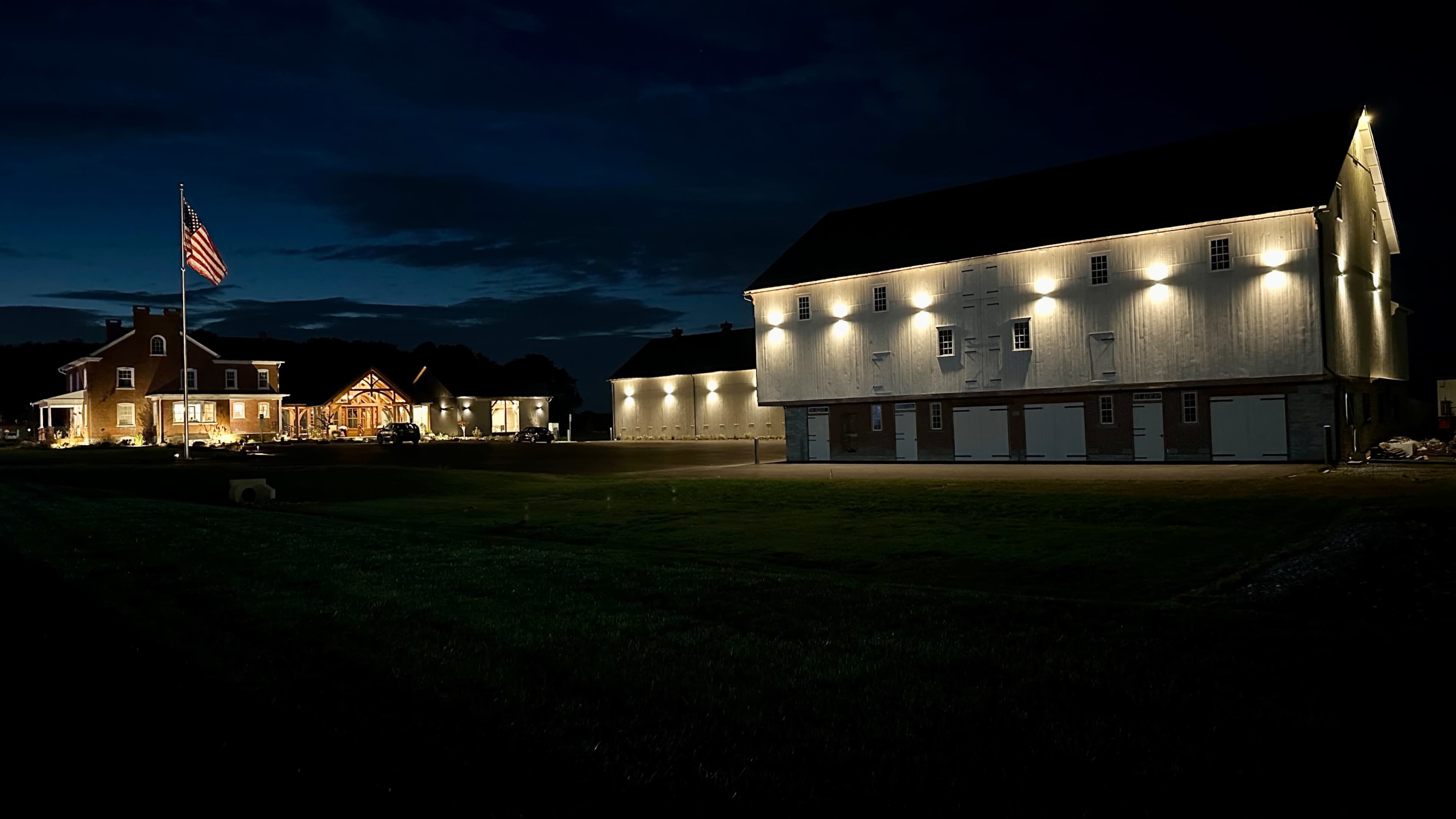 A well-lit farmstead features a large barn and a house, with an American flag prominently displayed, under a dark sky.