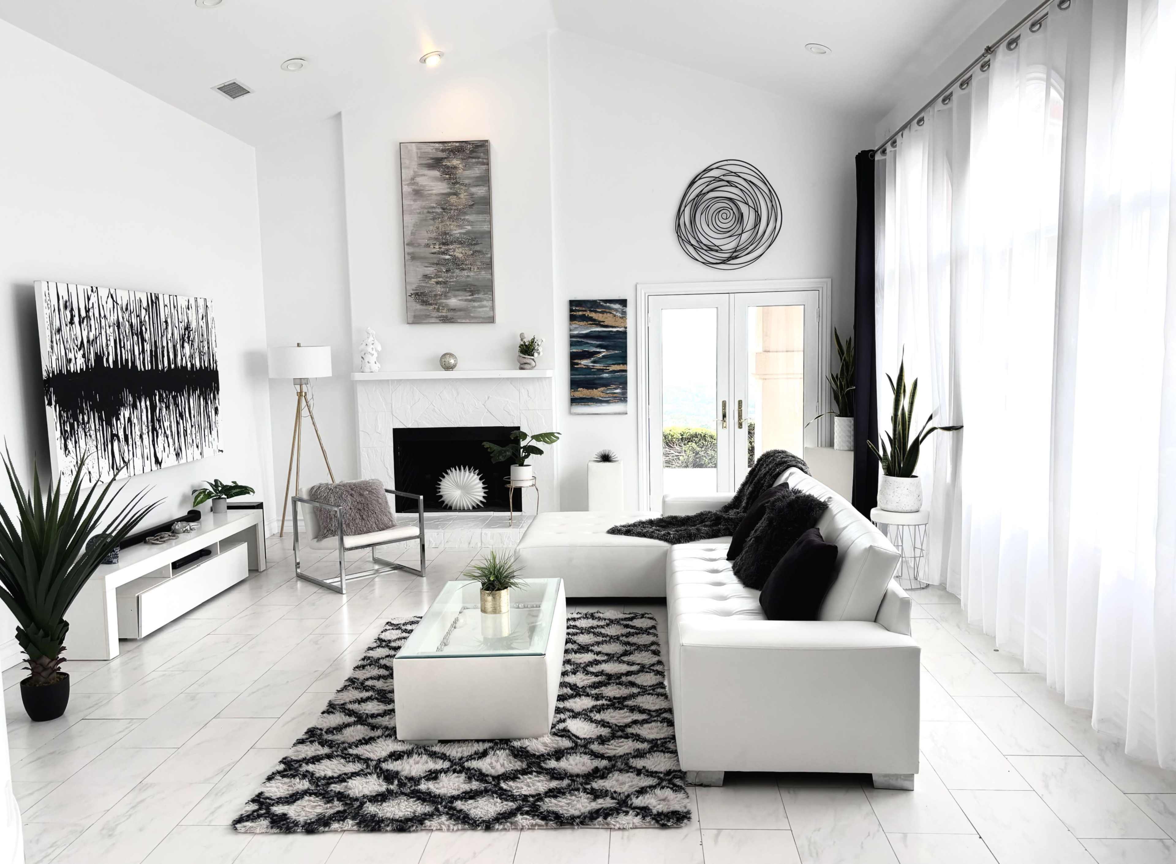 The image shows a modern living room featuring a white sectional sofa, a glass coffee table, potted plants, and large windows with sheer curtains.