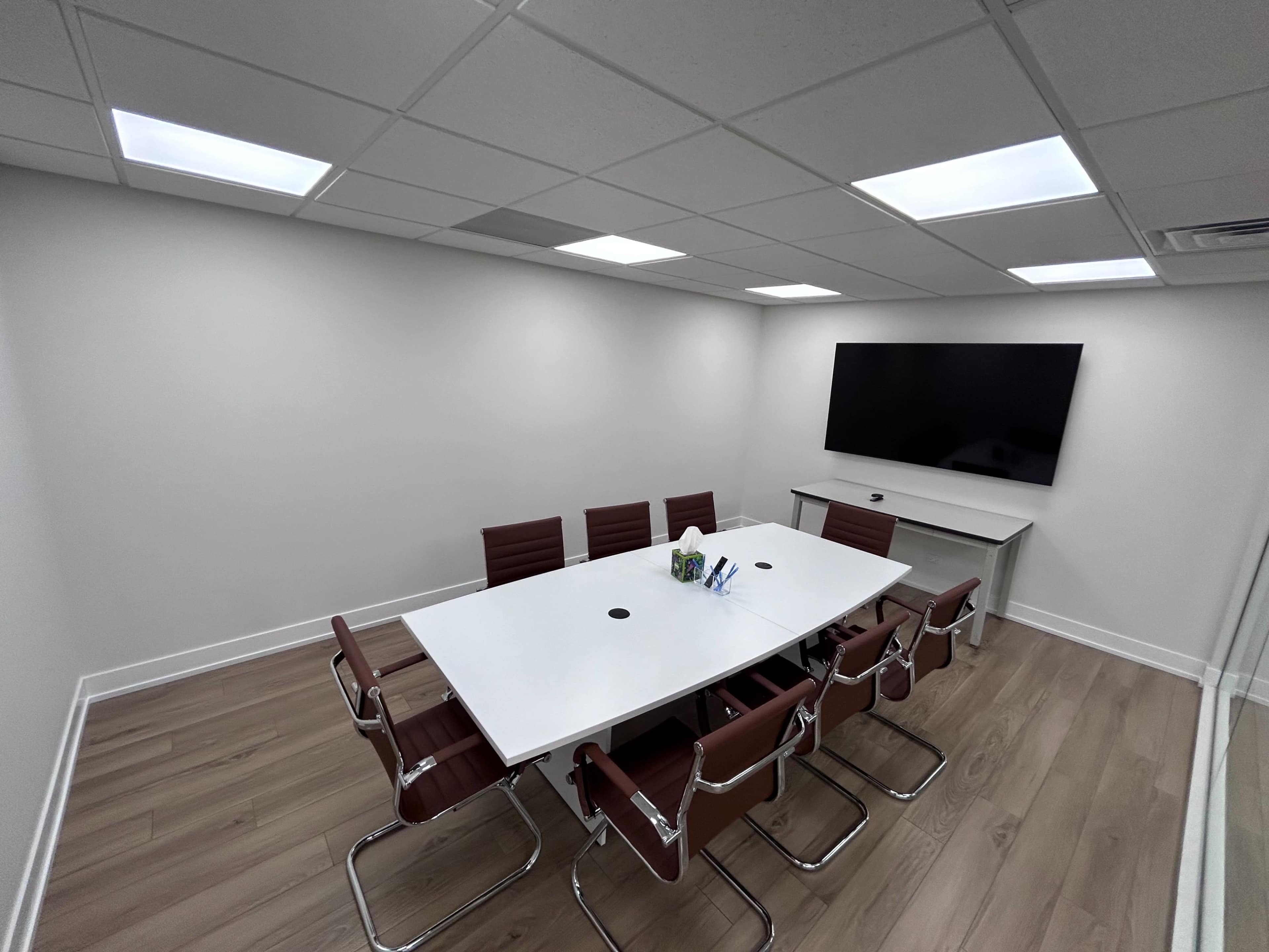 The image shows a modern conference room with a large white table, eight brown leather chairs, and a wall-mounted TV.