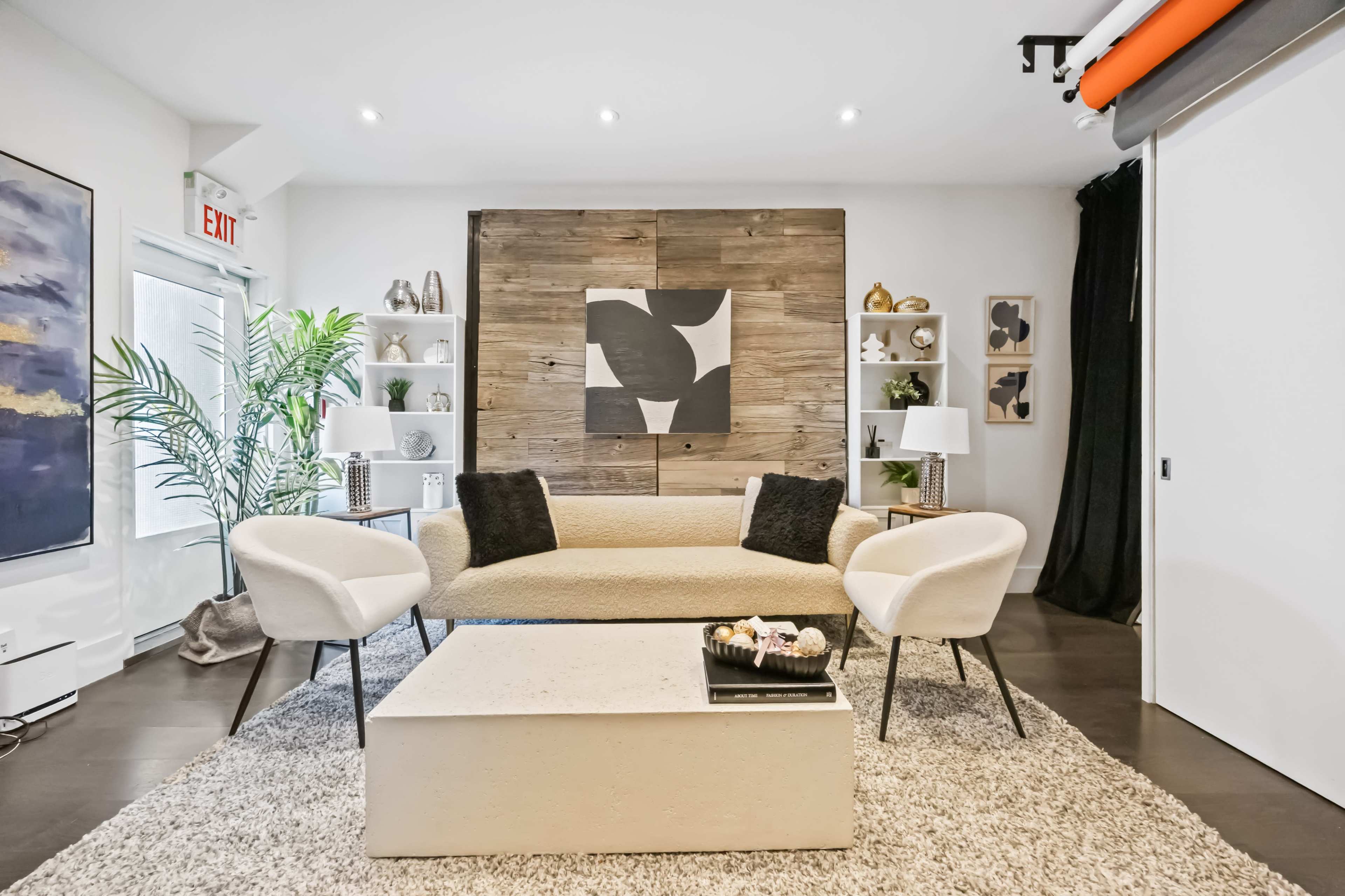 The image depicts a modern living room featuring a beige sofa, two white accent chairs, and a light-colored coffee table, all set against a backdrop of wood paneling and decorative shelving.