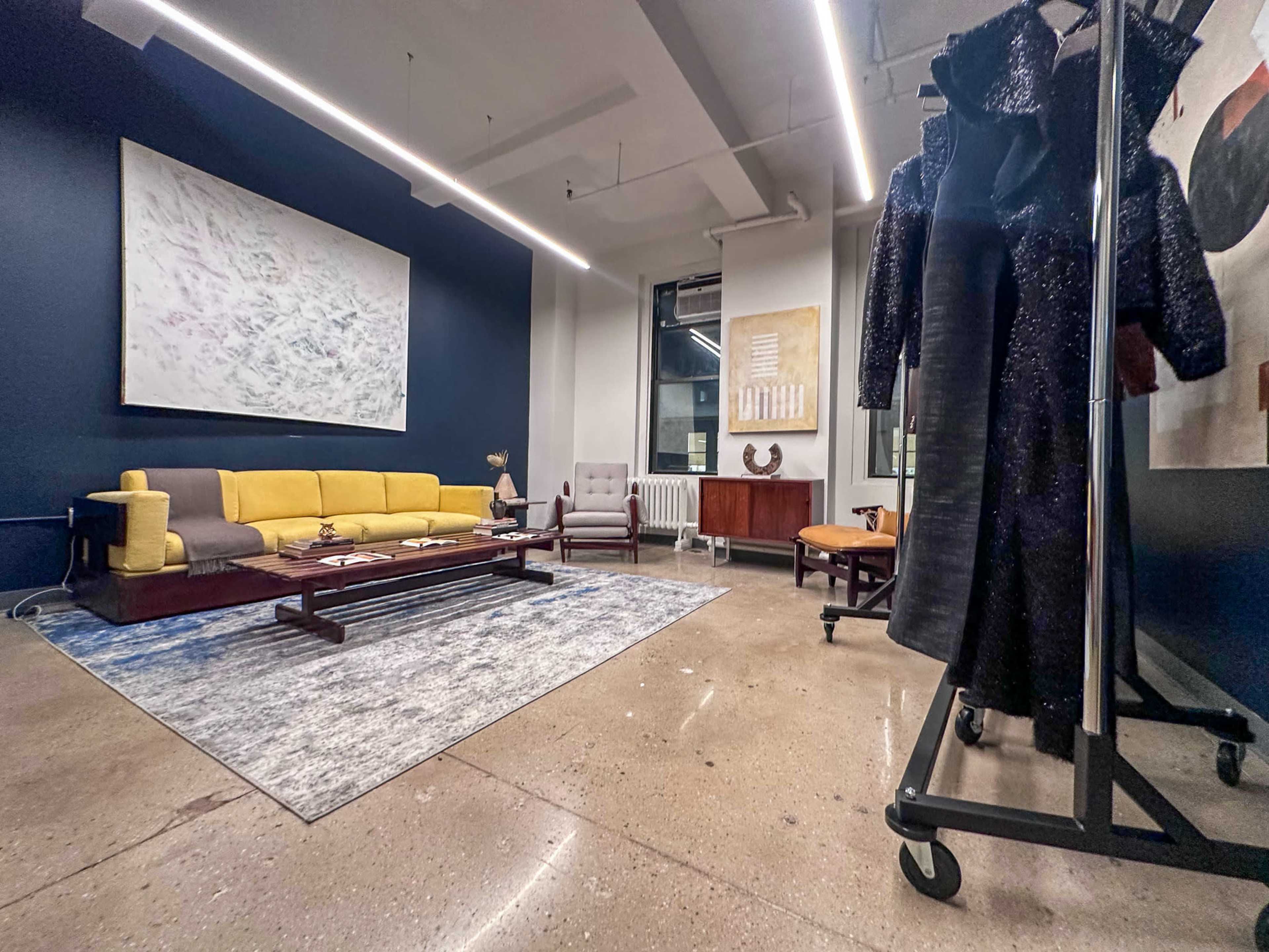 The image shows a spacious modern interior featuring a yellow sofa, a gray armchair, a wooden coffee table, and art pieces on the walls, with a clothing rack on one side.