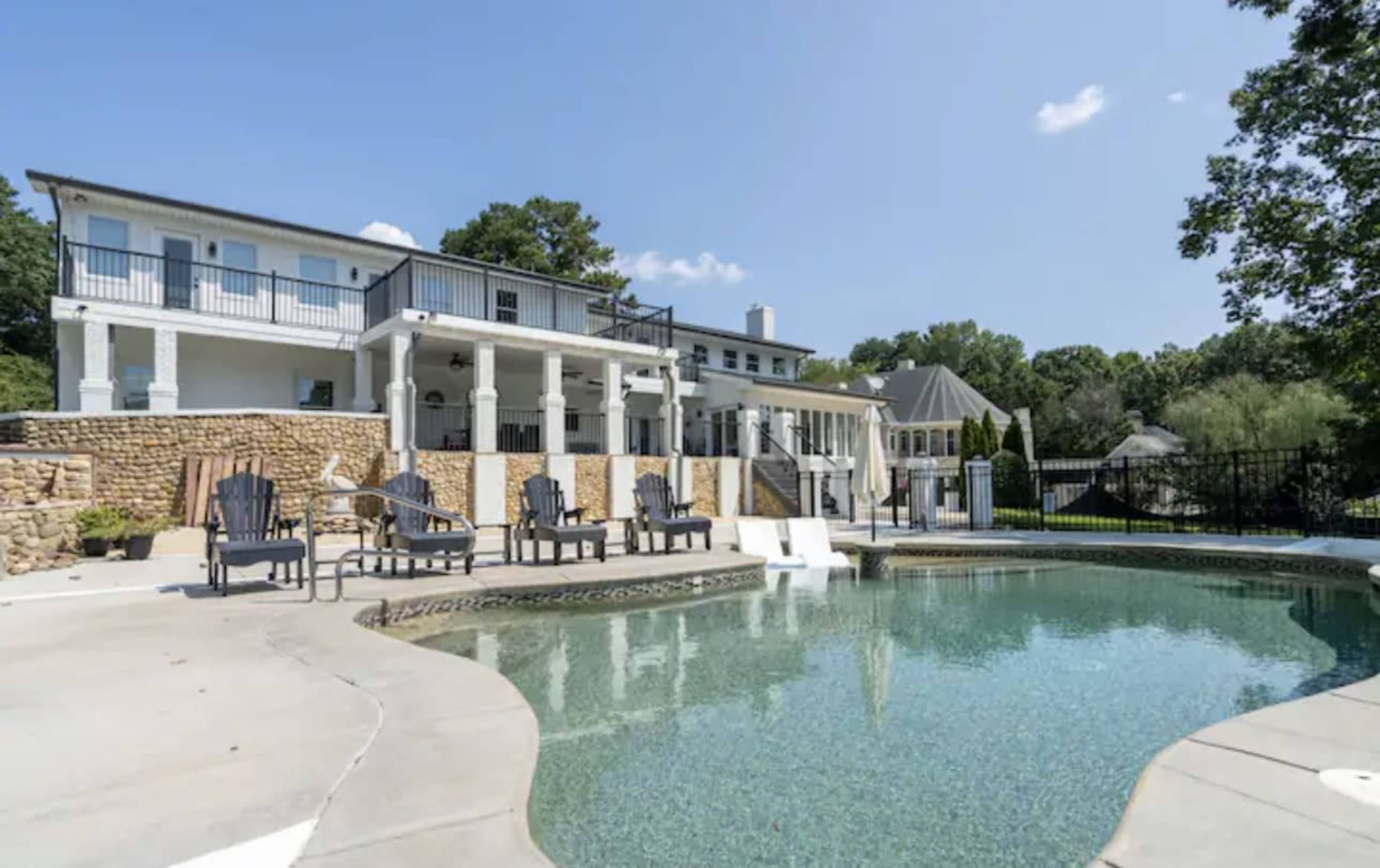 A large, two-story white house with a stone wall overlooks a swimming pool surrounded by adirondack chairs.