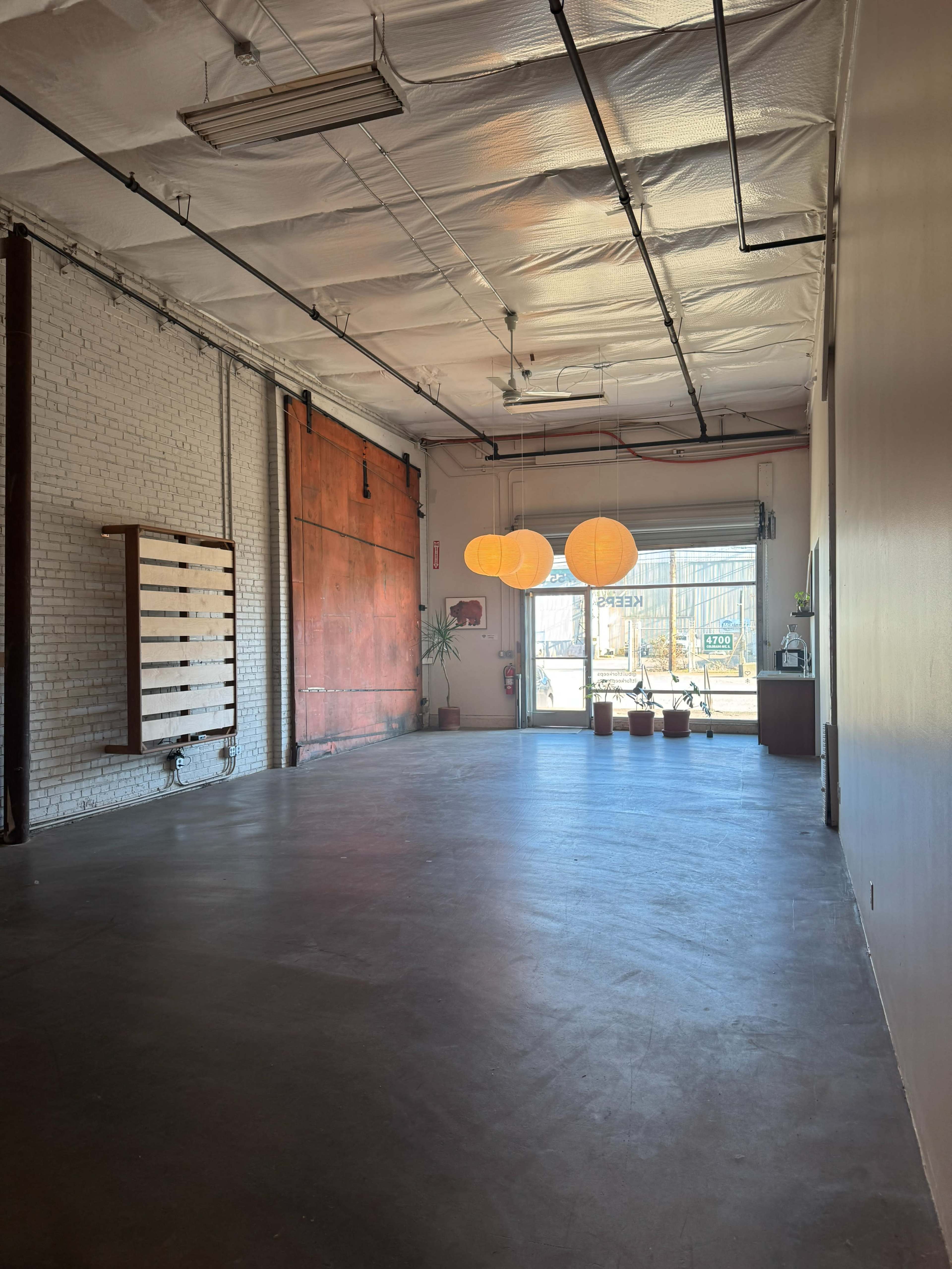 The image shows an empty interior space with a concrete floor, exposed brick walls, and three hanging orange lanterns.