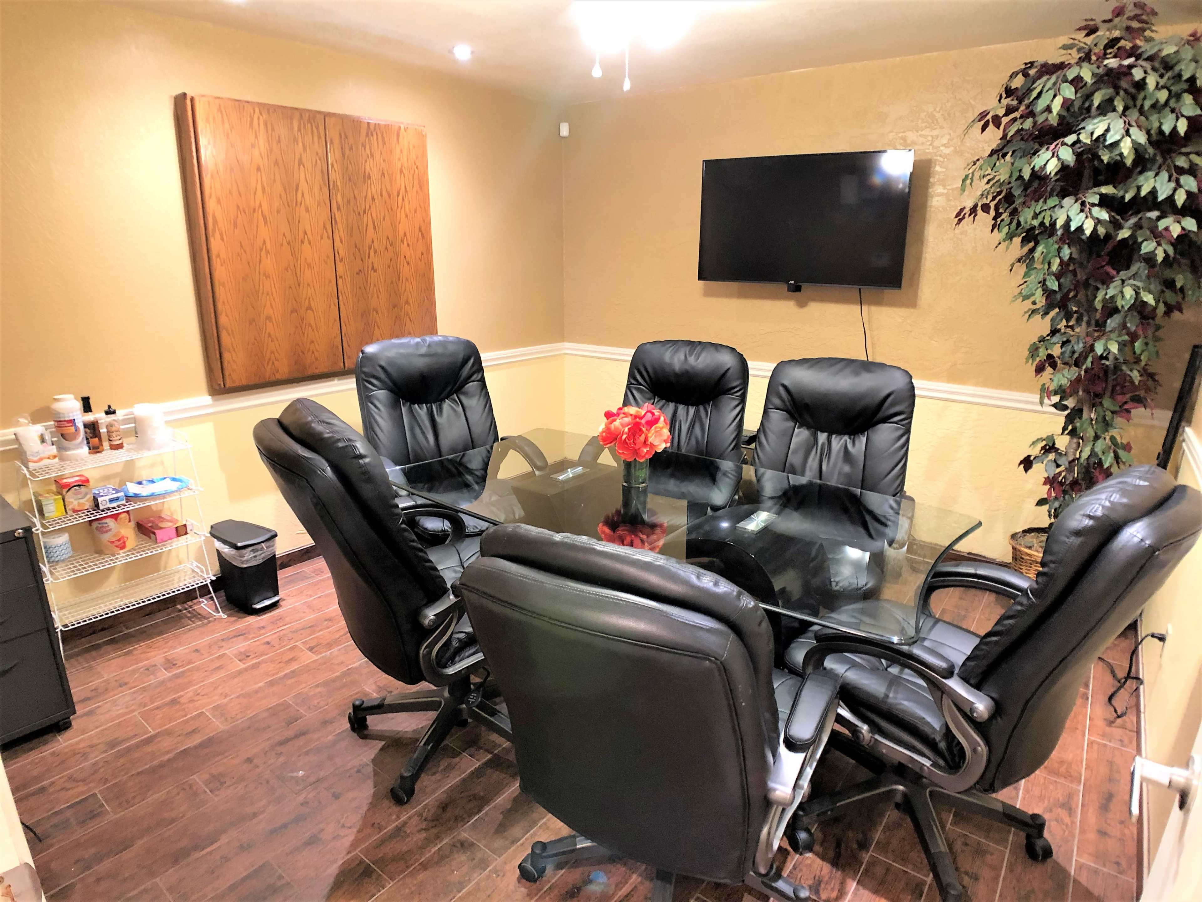 The image shows a meeting room with a glass conference table surrounded by six black leather chairs, a television on the wall, and a decorative plant in the corner.