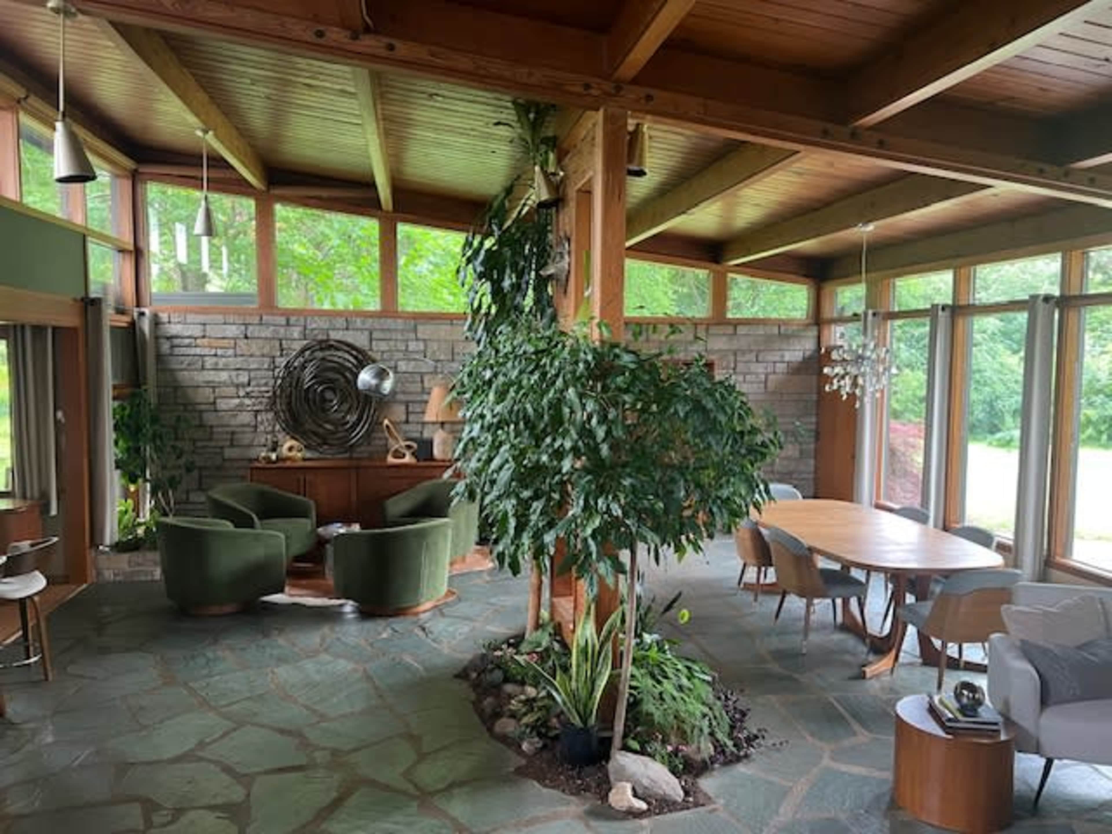 The interior of a spacious, modern living area features large windows, wooden beams, a stone wall, and a mix of green furnishings and plants.