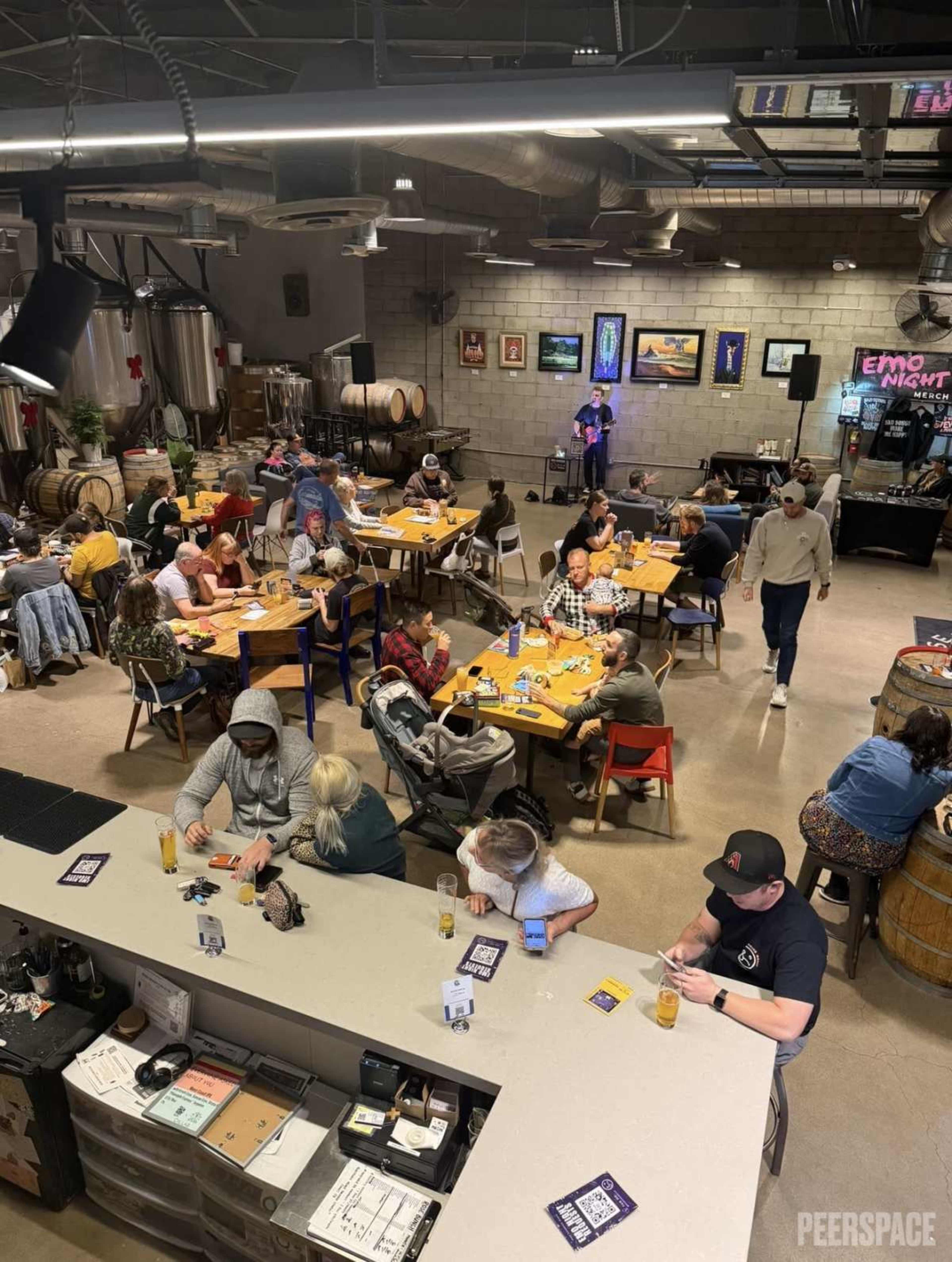 A brewery holds a lively gathering with groups of people seated at tables, enjoying food and drinks amidst large brewing equipment and barrels.