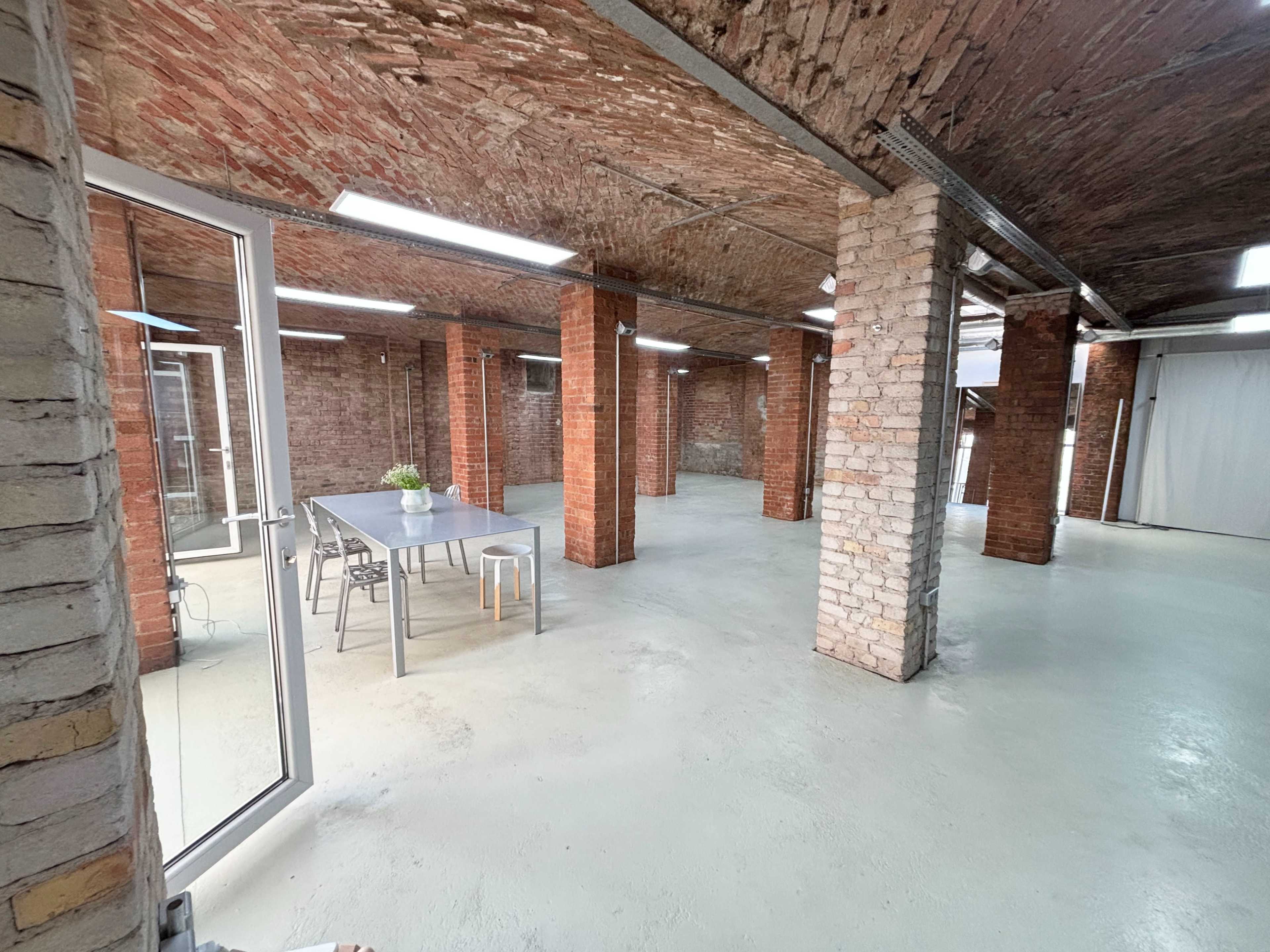 The image shows a large, open industrial space with exposed brick walls, a concrete floor, and a long table with chairs in the center.