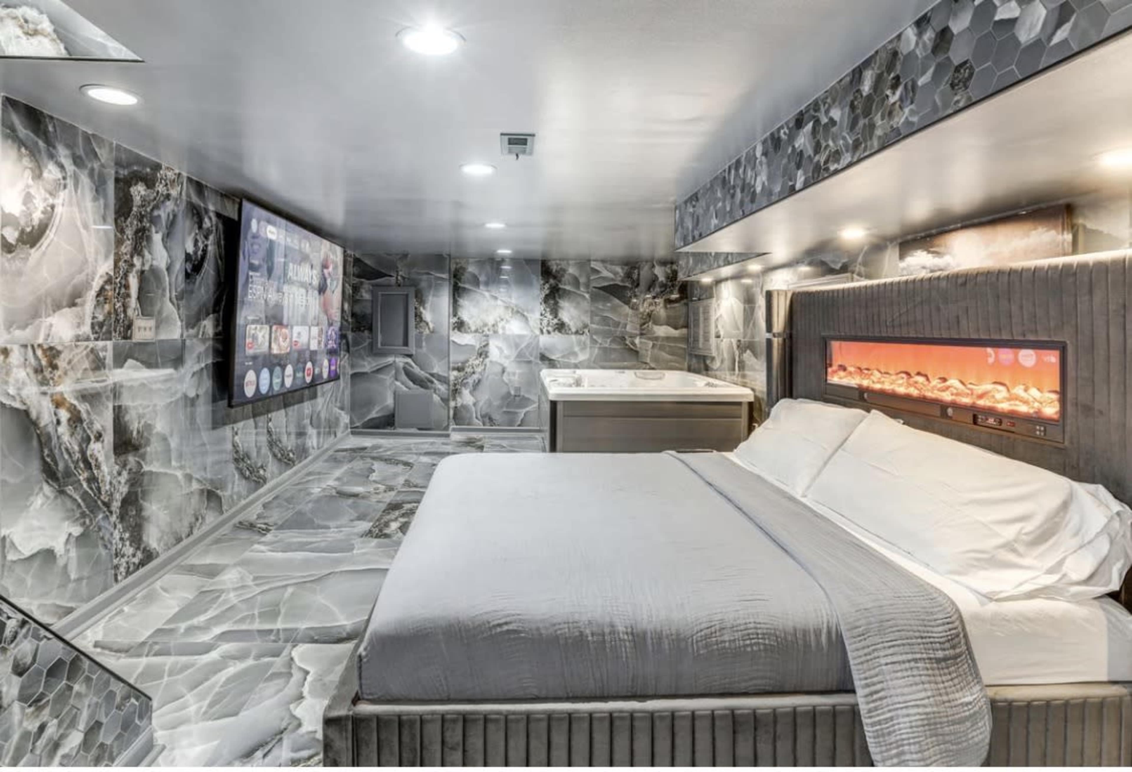 The image shows a modern bedroom with sleek, stone-like walls and flooring, featuring a large bed and a wall-mounted TV.