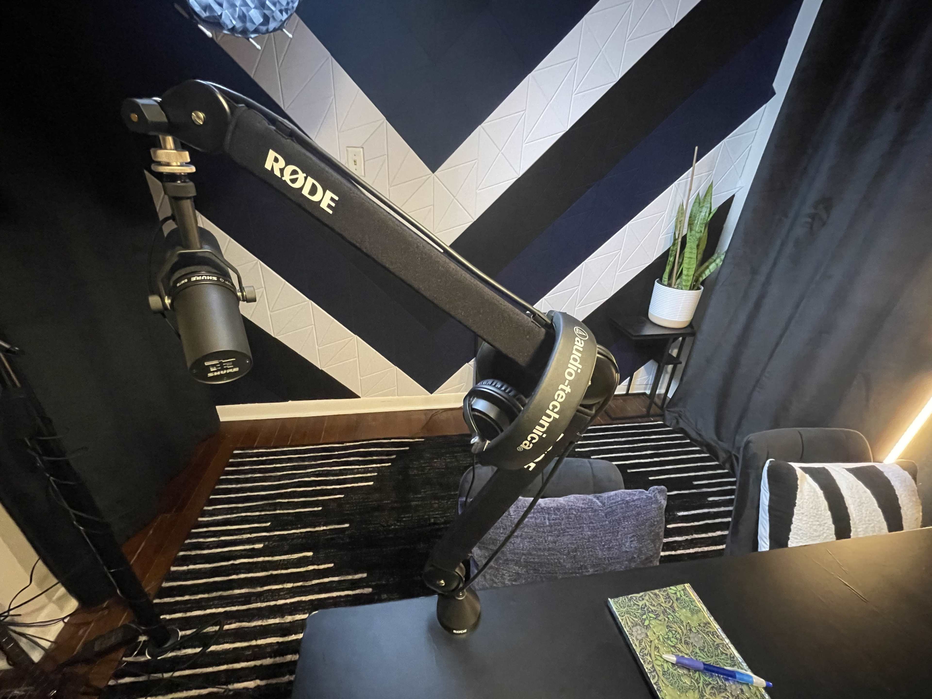 A black microphone mounted on an adjustable arm is positioned on a desk in a room with geometric patterned walls and a small plant.