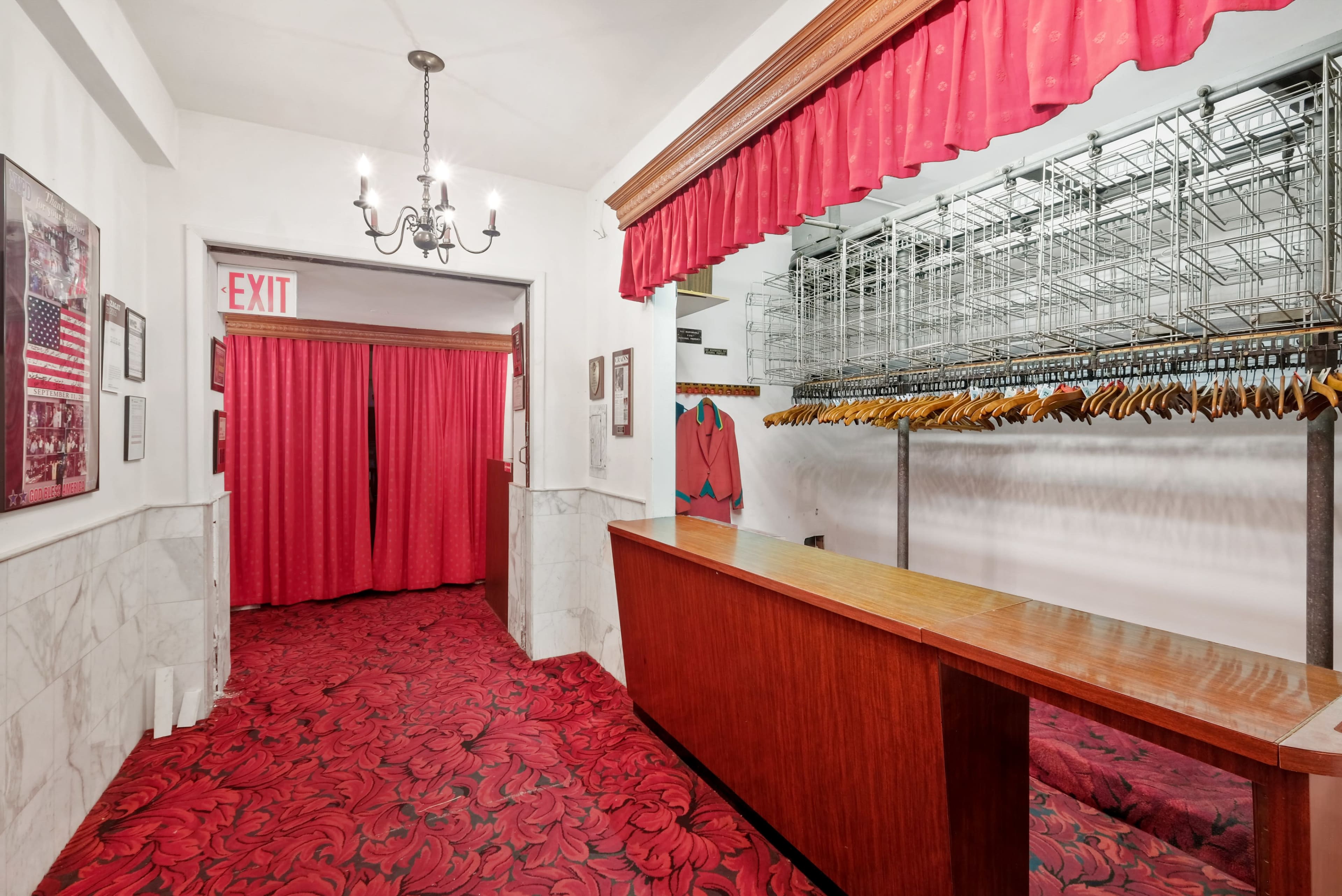 A hallway with a red carpet, a wooden reception desk, and red curtains leading to another area, alongside a row of coat hangers on a rack.